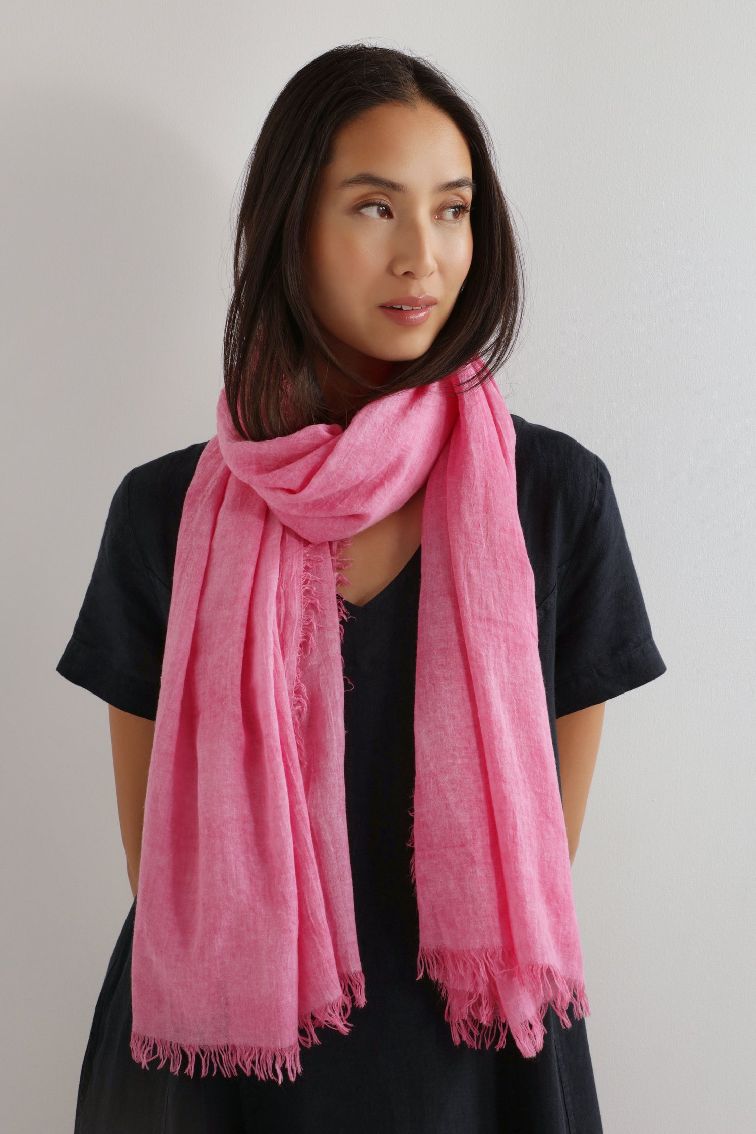 A woman with long dark hair and a black outfit stands by a light-colored wall, styled with the Soft Fringe Scarf in bright pink. The scarf’s textured material adds vibrant color and versatile styling to her look.