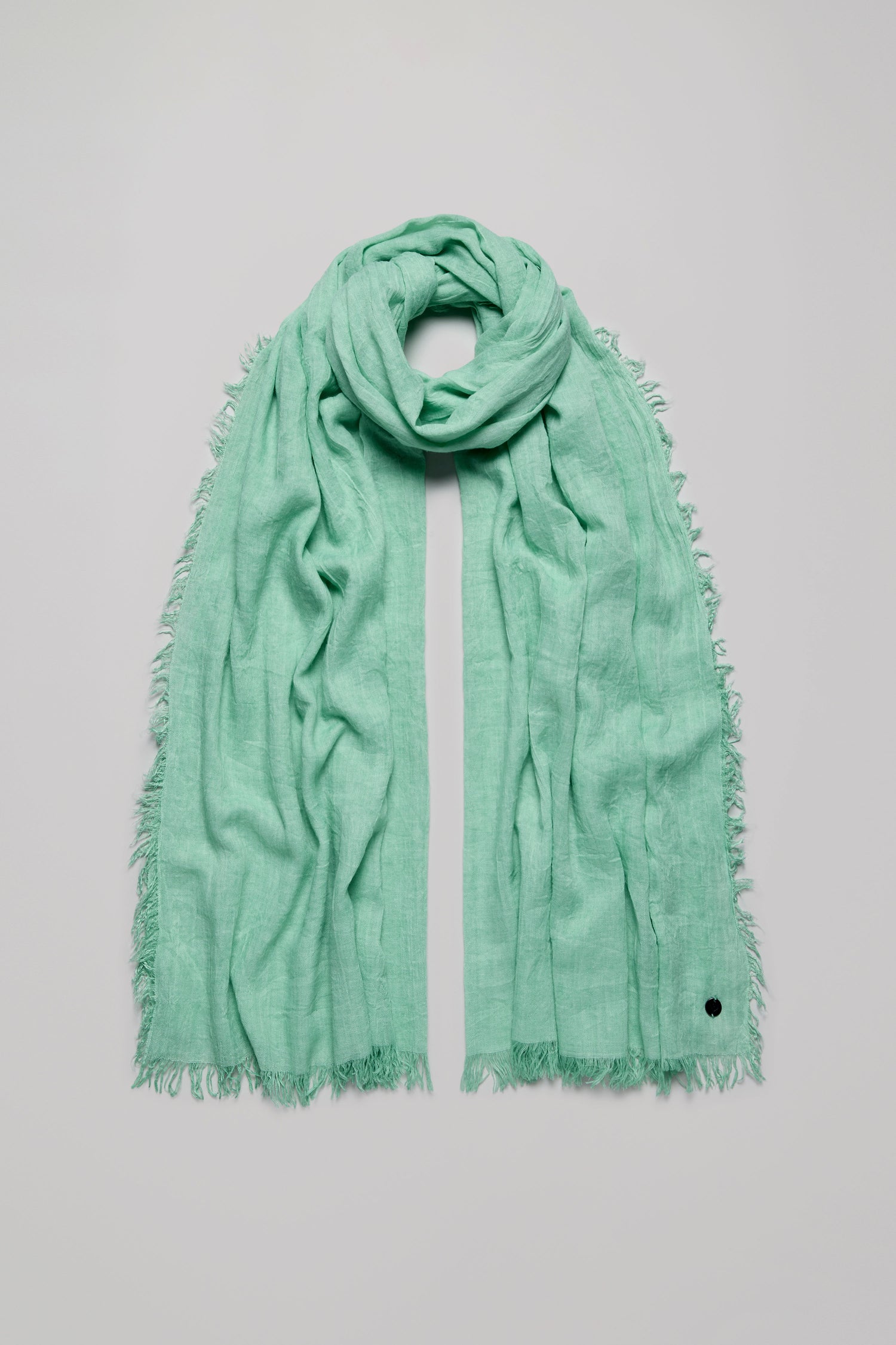 The Soft Fringe Scarf in light green features a textured material and frayed edges, shown loosely looped on a plain light background.