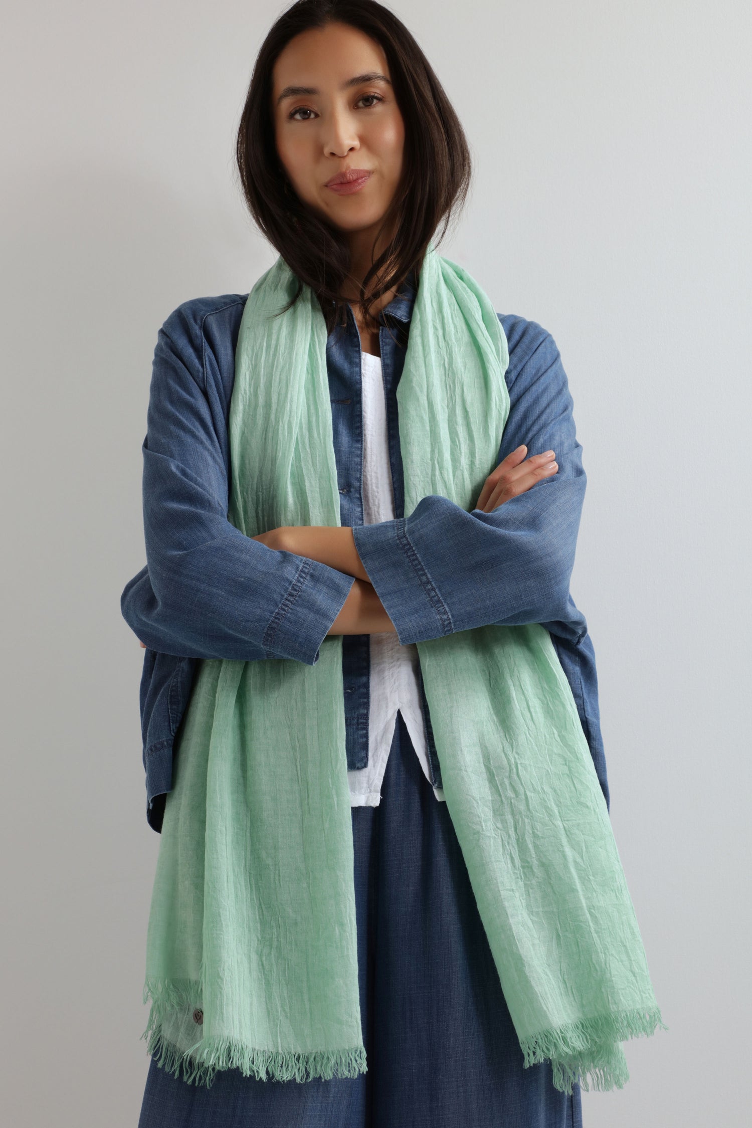 A woman with long dark hair stands with crossed arms, wearing a blue shirt over a white top and the Soft Fringe Scarf in a light green textured fabric, stylishly showcasing its versatility against a plain light background.