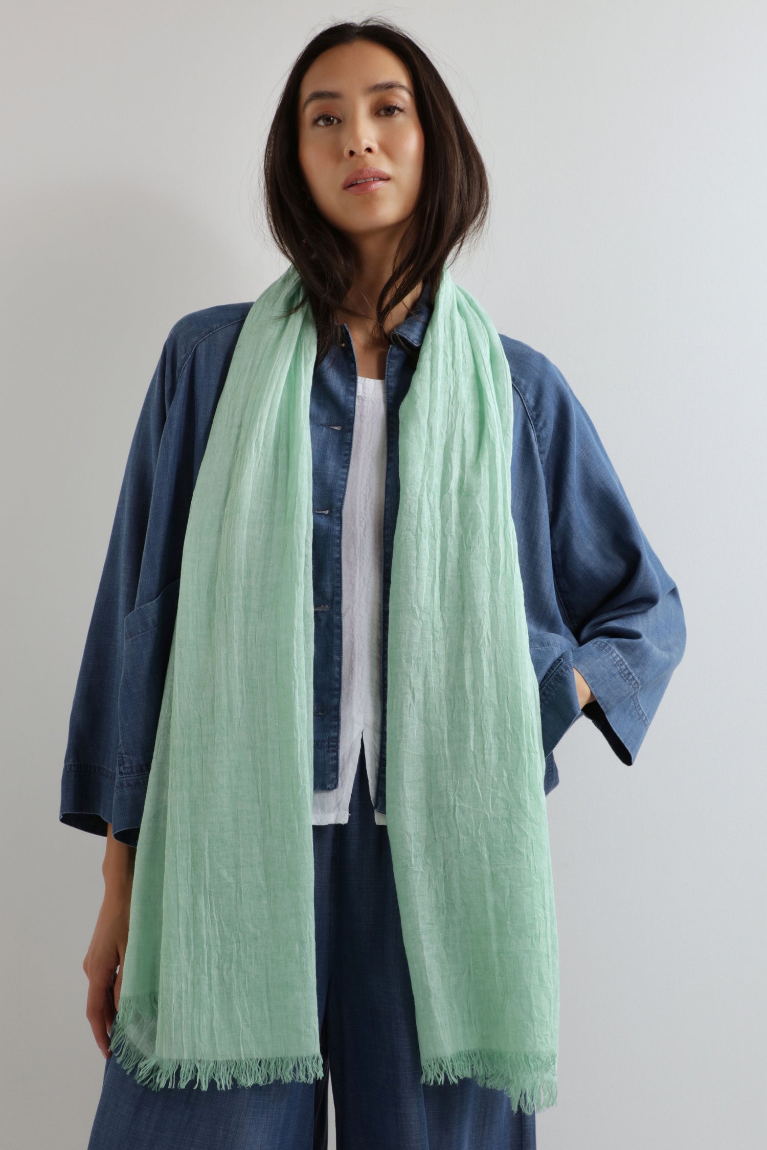 A woman stands against a plain light background wearing a loose blue shirt and pants, with the Soft Fringe Scarf in light green—crafted from textured material—draped around her neck.
