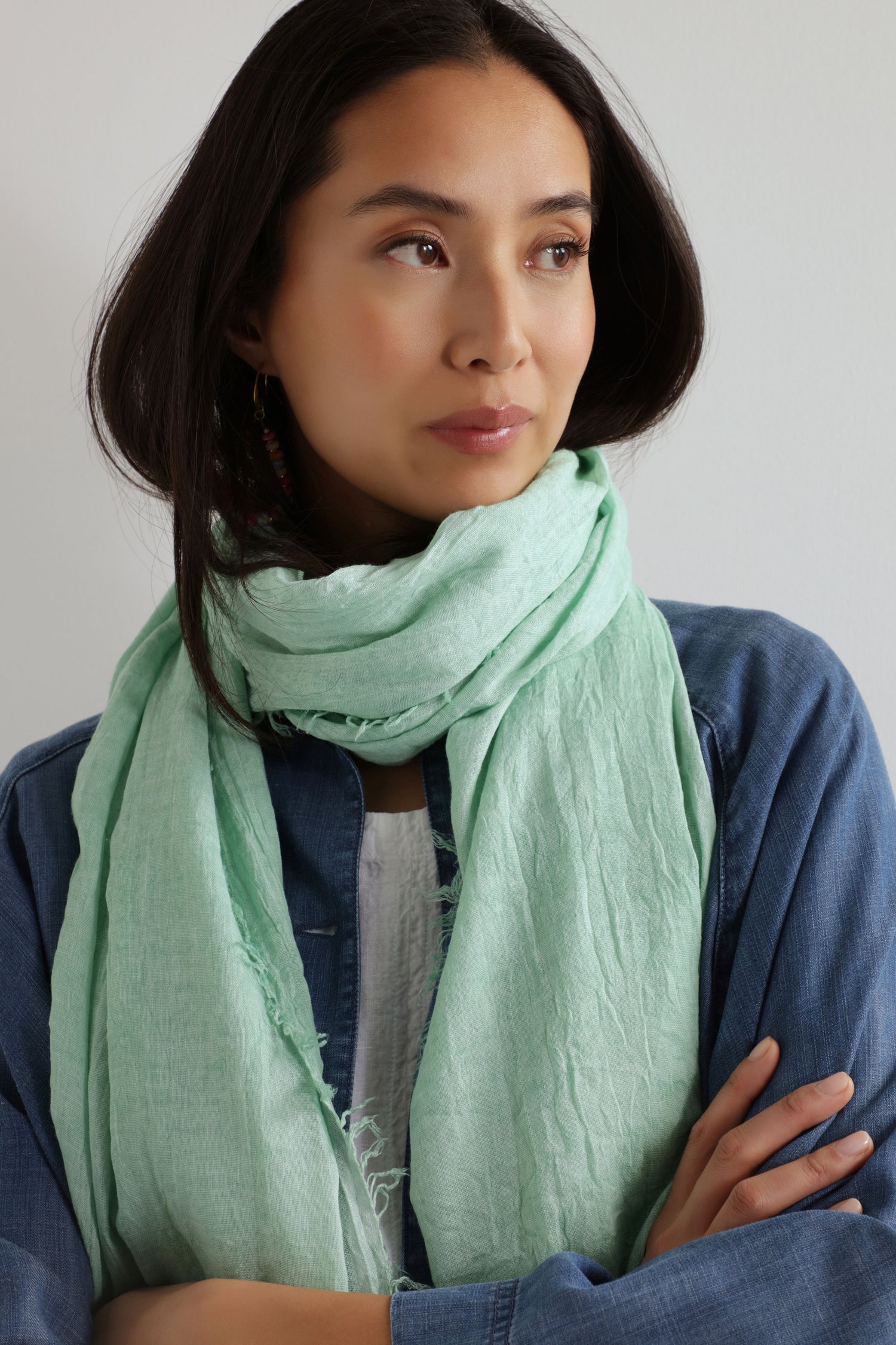 A woman with long dark hair and a denim shirt stands with arms crossed, modeling the Soft Fringe Scarf in light green. The scarf’s textured material adds visual interest for versatile styling against a plain background.