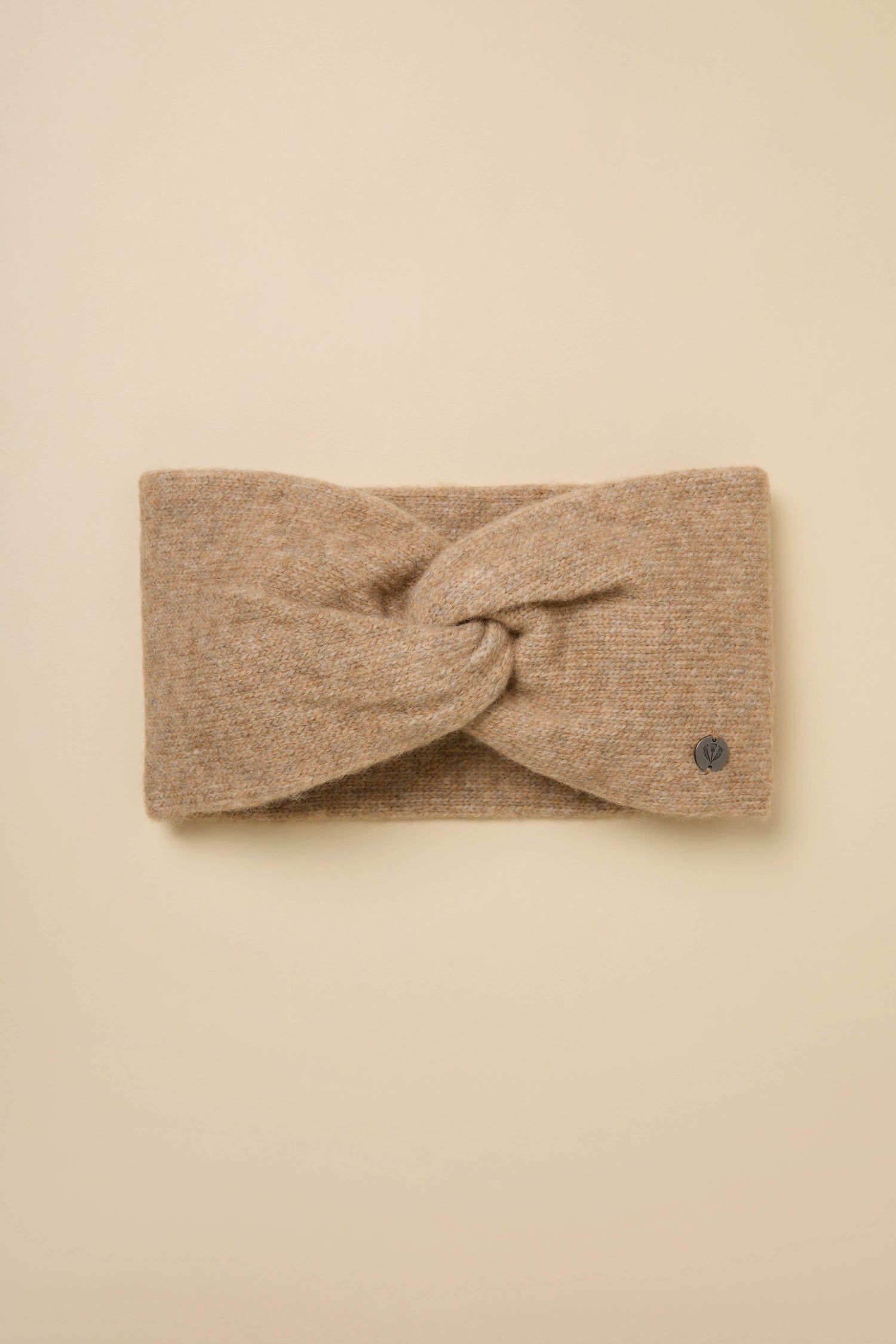 Wool Mix Sand Headband: Tan wool-blend knit with a twisted front and small round metal tag, shown laid flat on a beige background.