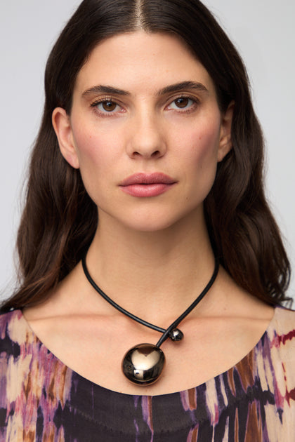 A woman with long dark hair in a patterned top wears the Charcoal Handmade Ceramic Bead Necklace—featuring a black cord and large ceramic bead—facing forward against a plain background.