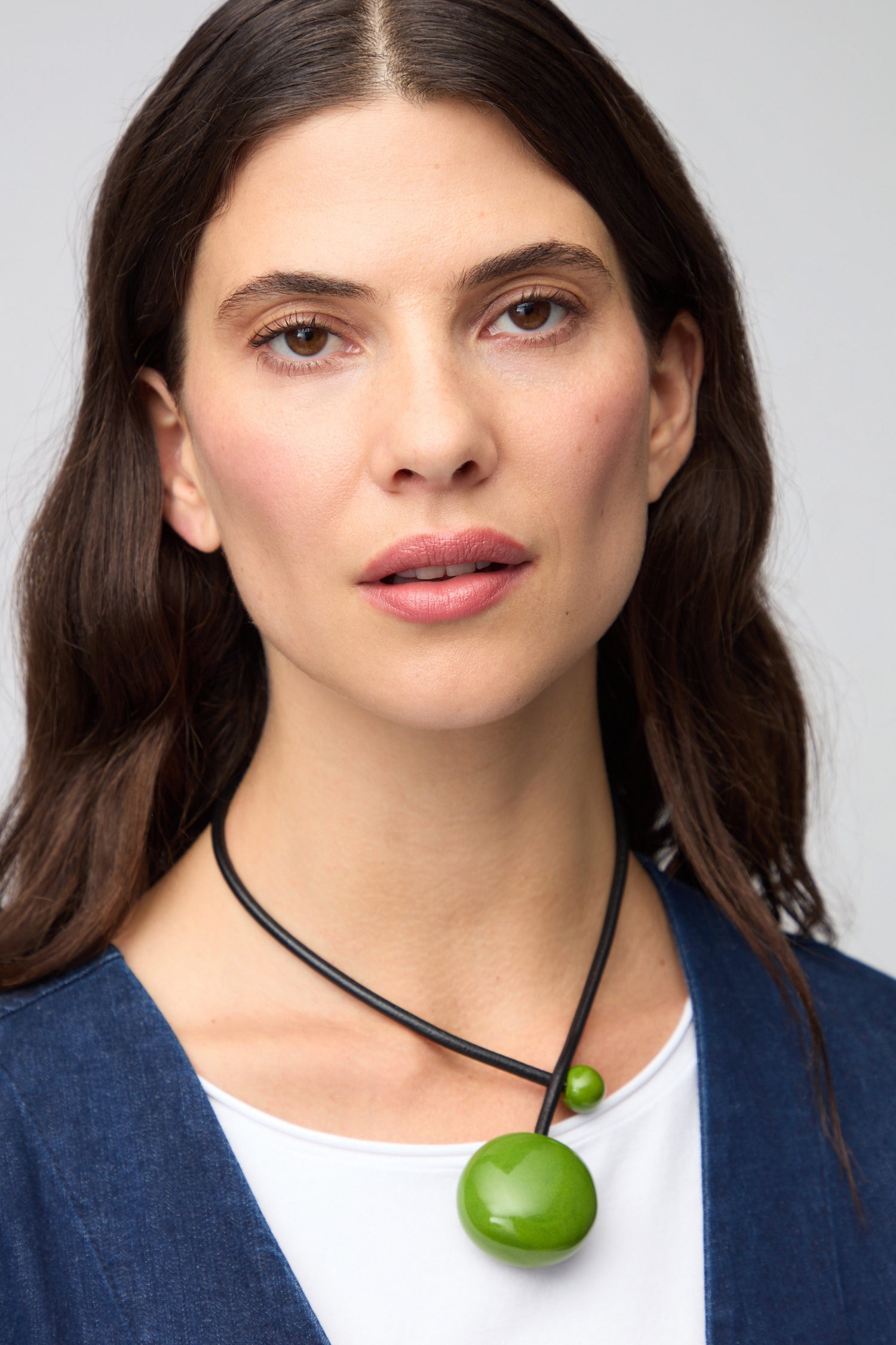 A woman with long brown hair, dressed in a blue top and white shirt, poses against a neutral background while accessorizing with the Moss Handmade Ceramic Bead Necklace.
