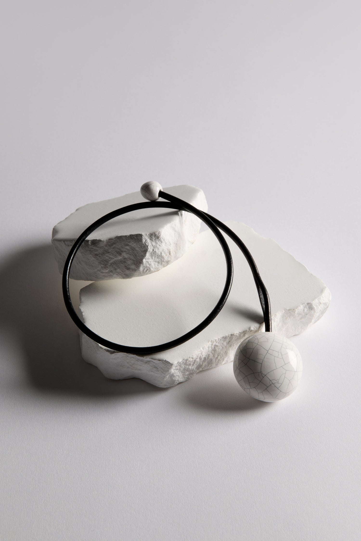 The Handmade Ceramic Crackle Bead Necklace features a cracked white ceramic bead on a black cord, displayed on textured white stone slabs.