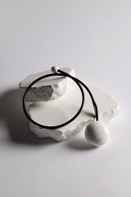 The Handmade Ceramic Crackle Bead Necklace features a cracked white ceramic bead on a black cord, displayed on textured white stone slabs.