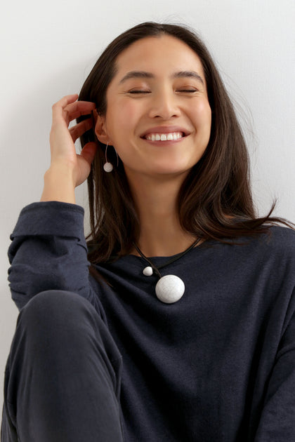 A woman with long dark hair smiles, wearing a dark top and the Handmade Ceramic Crackle Bead Necklace, with matching earrings, seated against a light background.