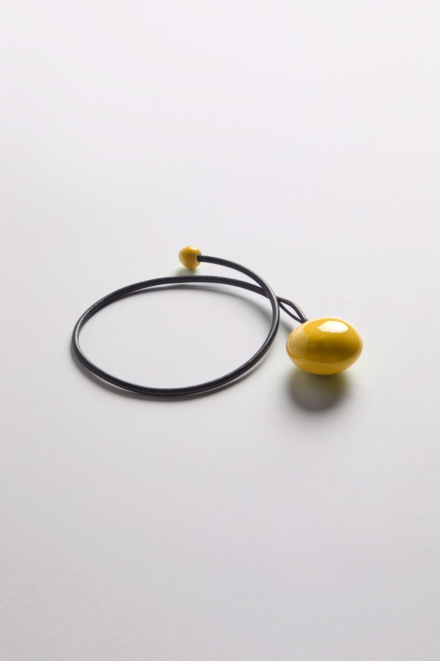 The Handmade Ceramic Bead Necklace features a black cord with a round yellow pendant and small yellow bead, showcasing an organic finish against a white background.