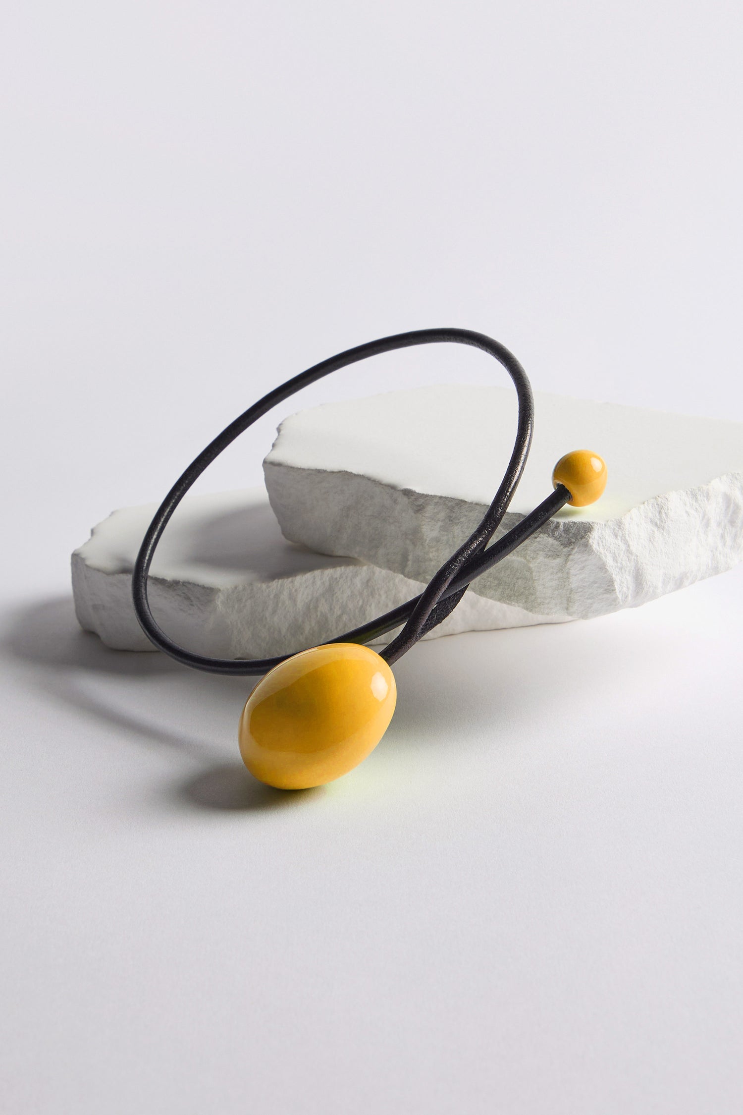 The Handmade Ceramic Bead Necklace features two yellow oval beads on a black cord with an organic finish, showcased on irregular white stone slabs against a plain background.