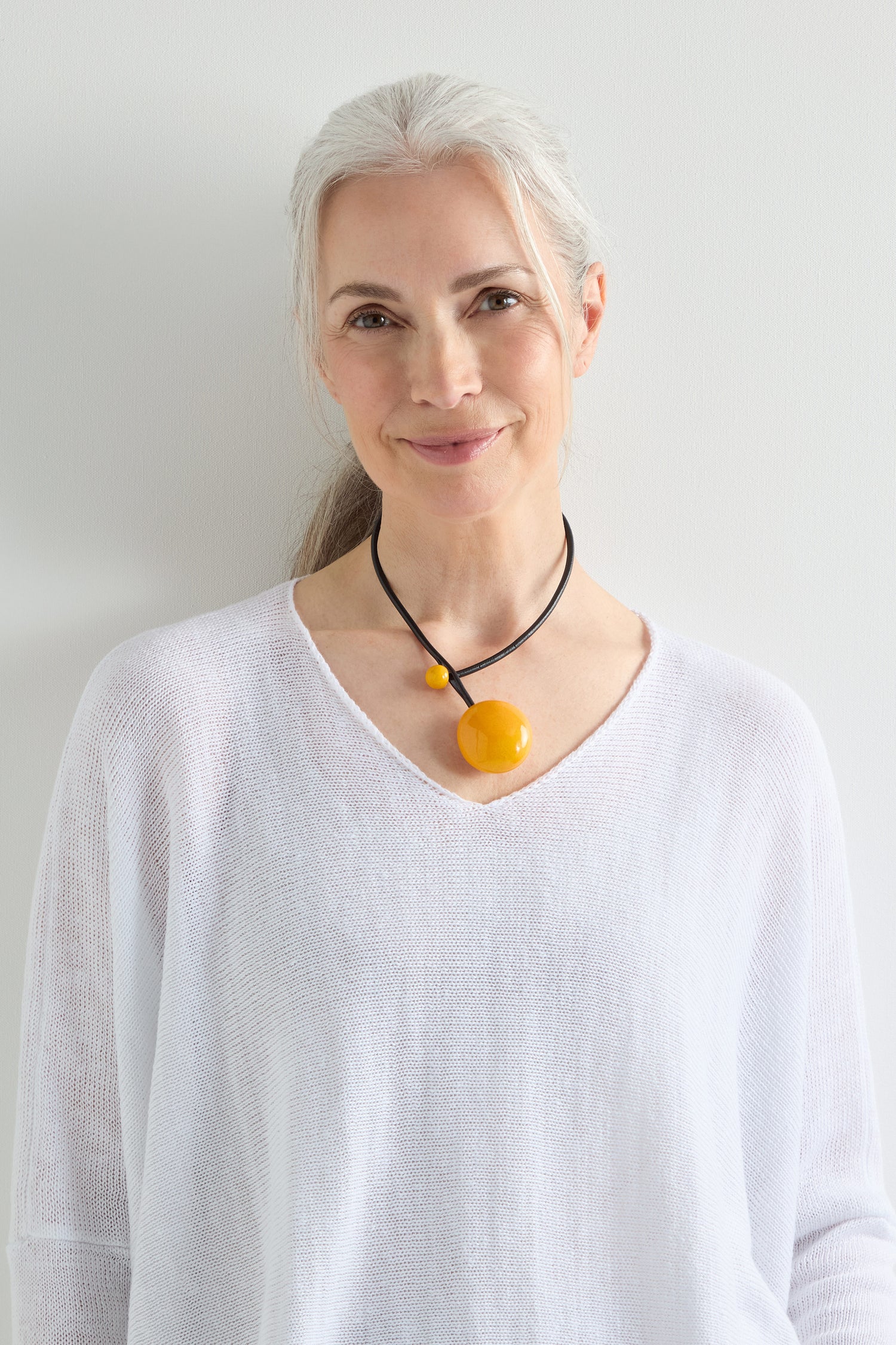 A woman with gray hair pulled back, wearing a white sweater and the Handmade Ceramic Bead Necklace featuring two yellow pendants, stands against a plain light-colored wall, smiling slightly.