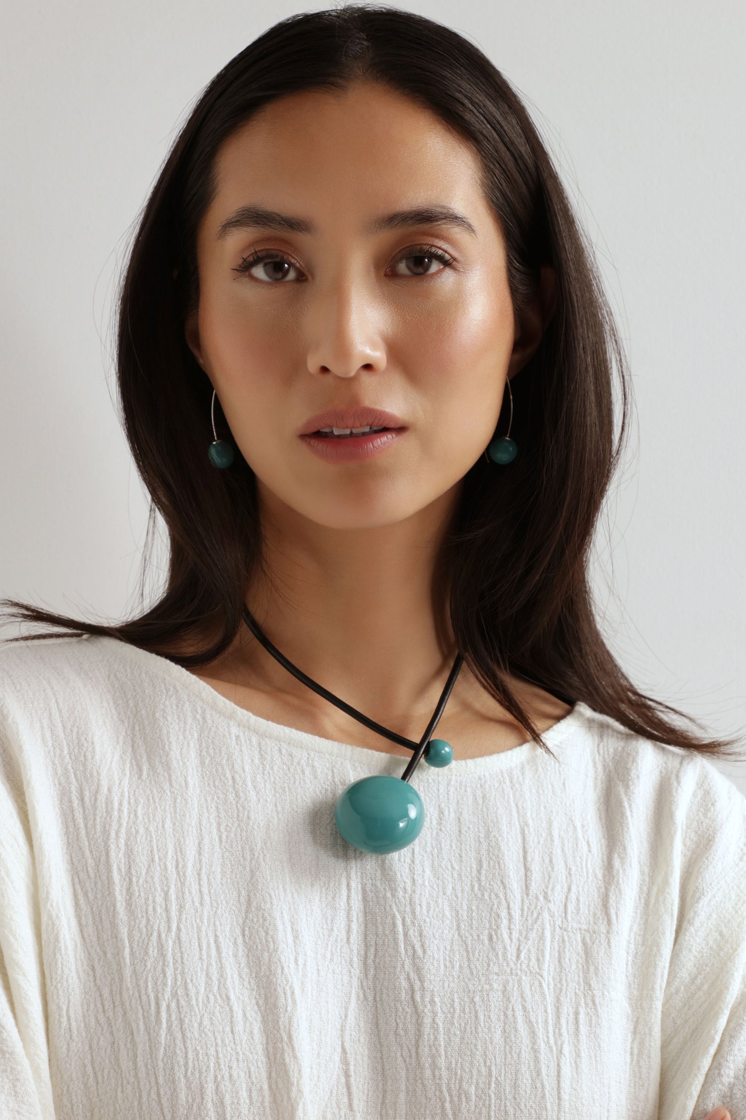 Woman with long dark hair wearing a white textured top and the Handmade Ceramic Bead Necklace, featuring an organic finish, stands against a plain light background.