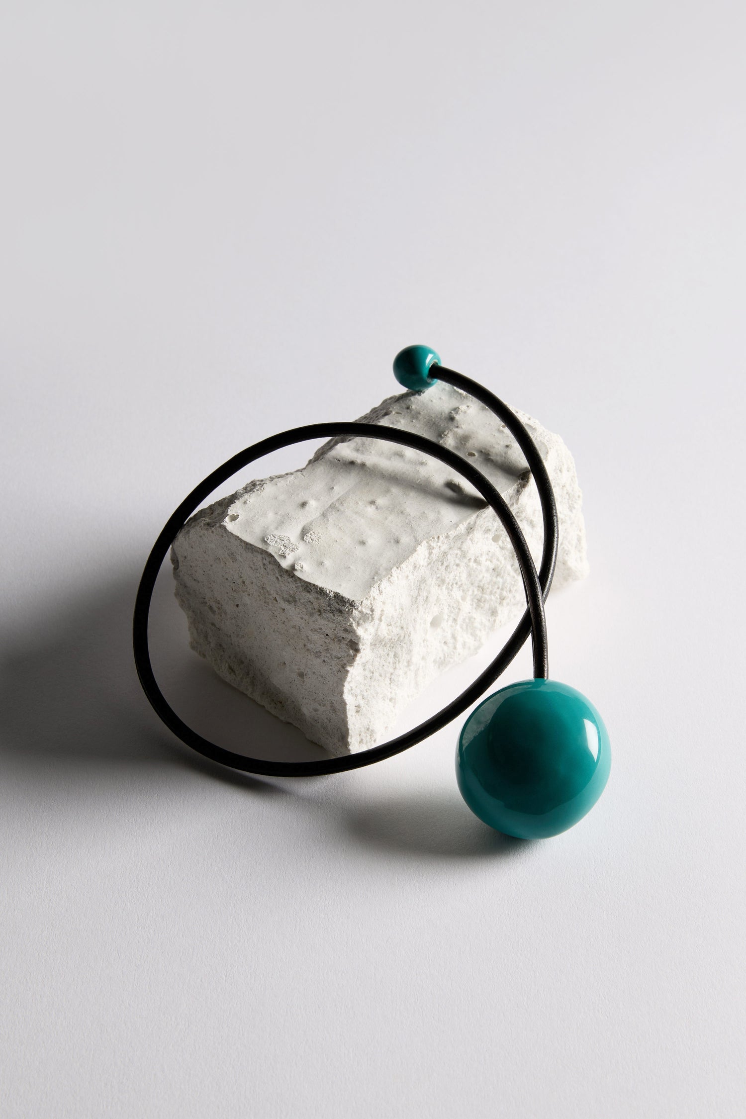 The Handmade Ceramic Bead Necklace, featuring a black cord with large and small teal ceramic beads in an organic finish, is displayed on a rough white rectangular stone block against a plain background.
