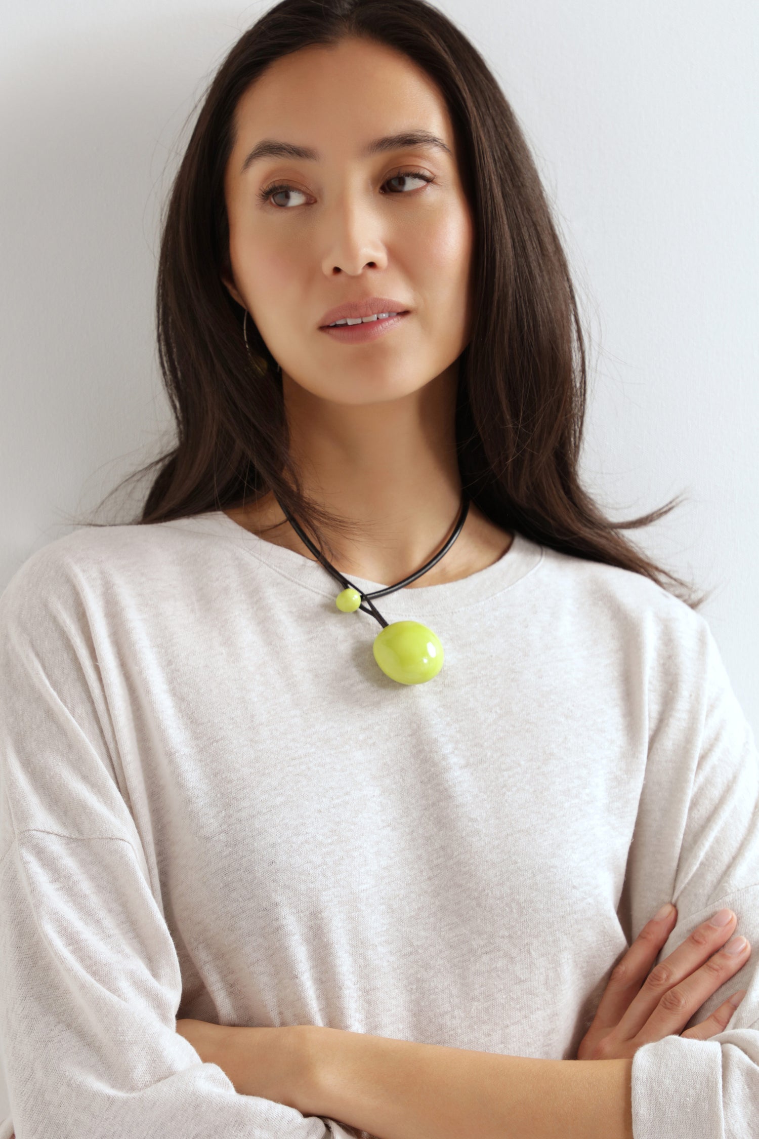 Woman with long dark hair wears a light beige top and a Handmade Ceramic Bead Necklace featuring a large, round green pendant, standing against a plain white wall with arms crossed.