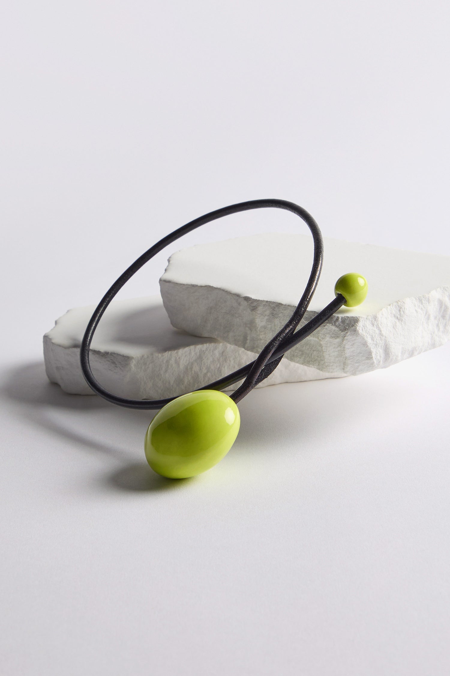 The Handmade Ceramic Bead Necklace features a black cord with large and small shiny lime-green oval beads, displayed on white stone slabs, with a sculptural twist that pairs perfectly with handmade ceramic bead earrings.