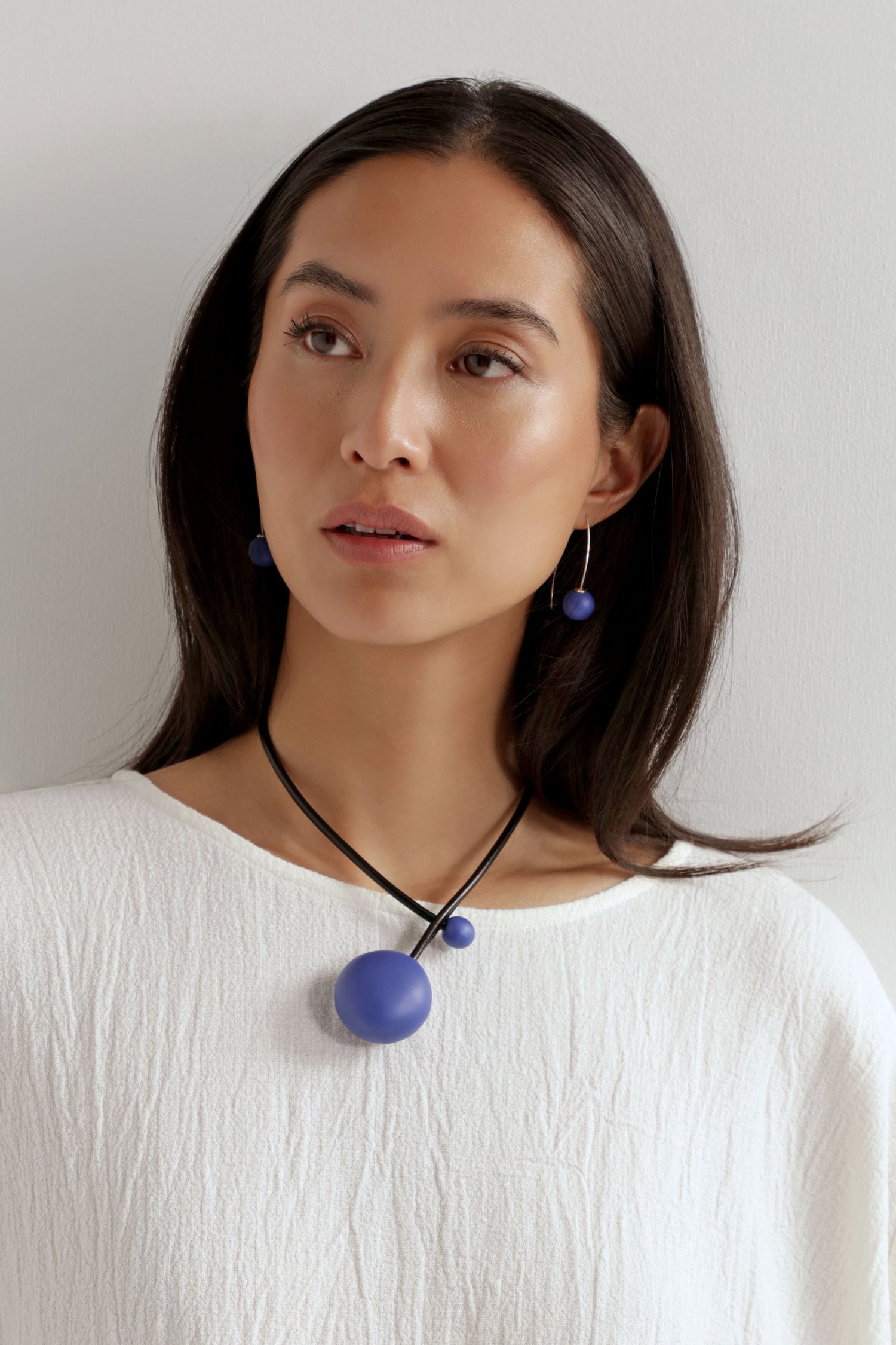 A woman with straight dark hair wears a textured white top and the Handmade Ceramic Bead Necklace, featuring two blue spheres, all set against a plain light background.