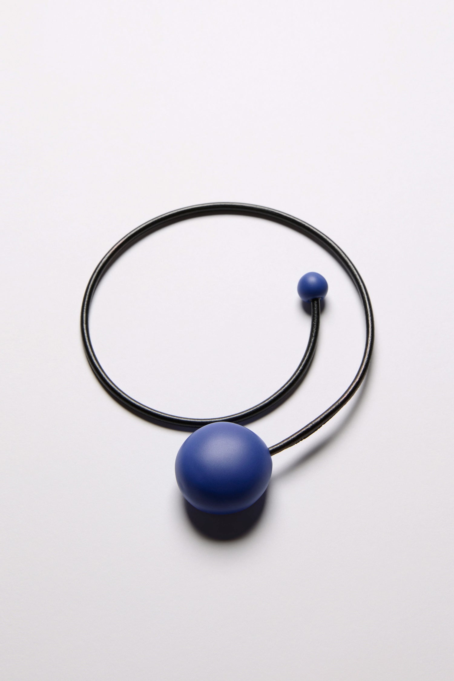 The Handmade Ceramic Bead Necklace features a thin black circular band with large and small blue ceramic spheres at each end, offering a sculptural twist for modern elegance.