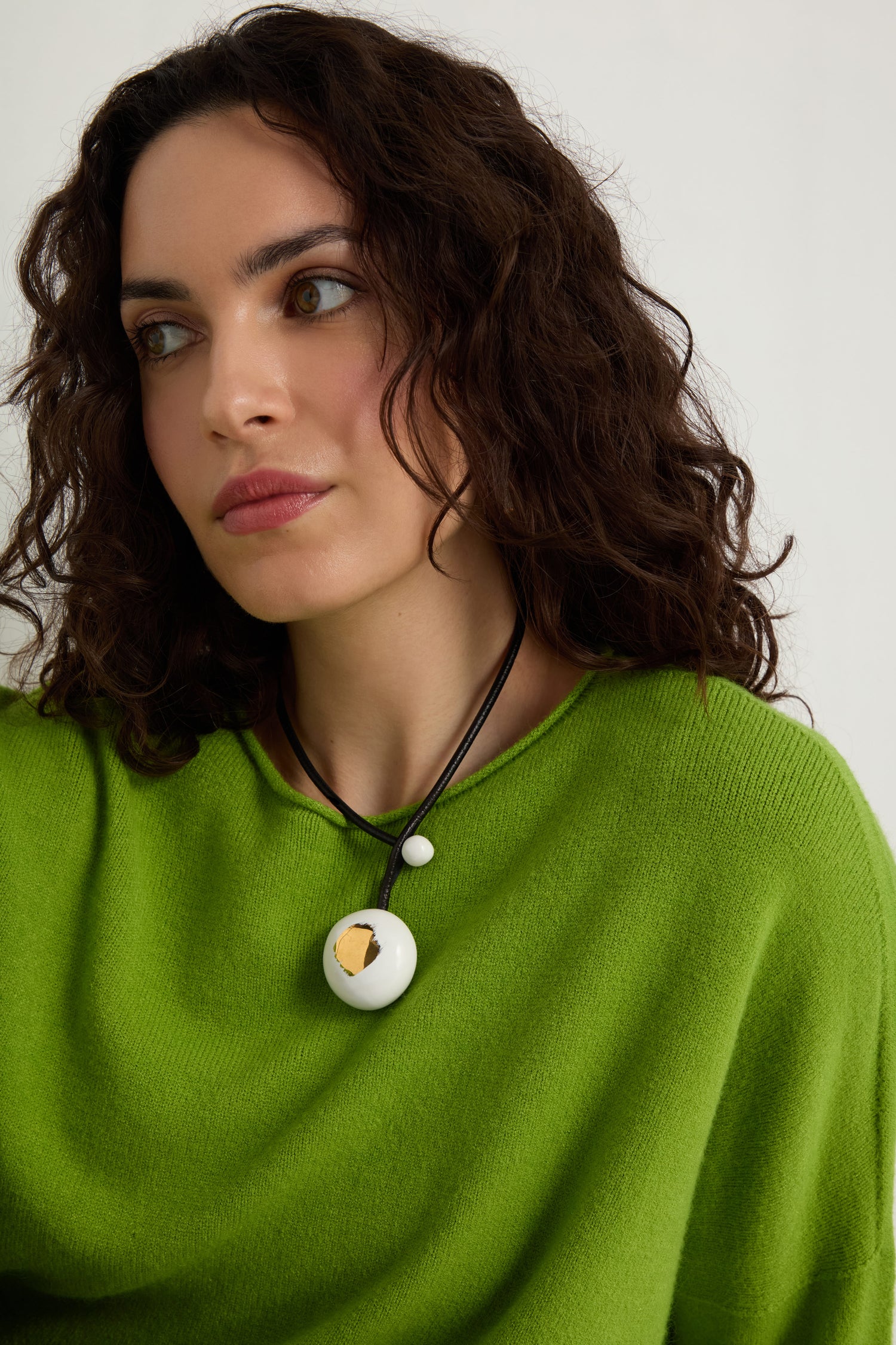 Woman with wavy dark hair in a bright green sweater wears the Handmade Gold Brushstroke Ceramic Bead Necklace—a black cord necklace featuring a round white ceramic bead pendant—while looking slightly to the side against a plain background.