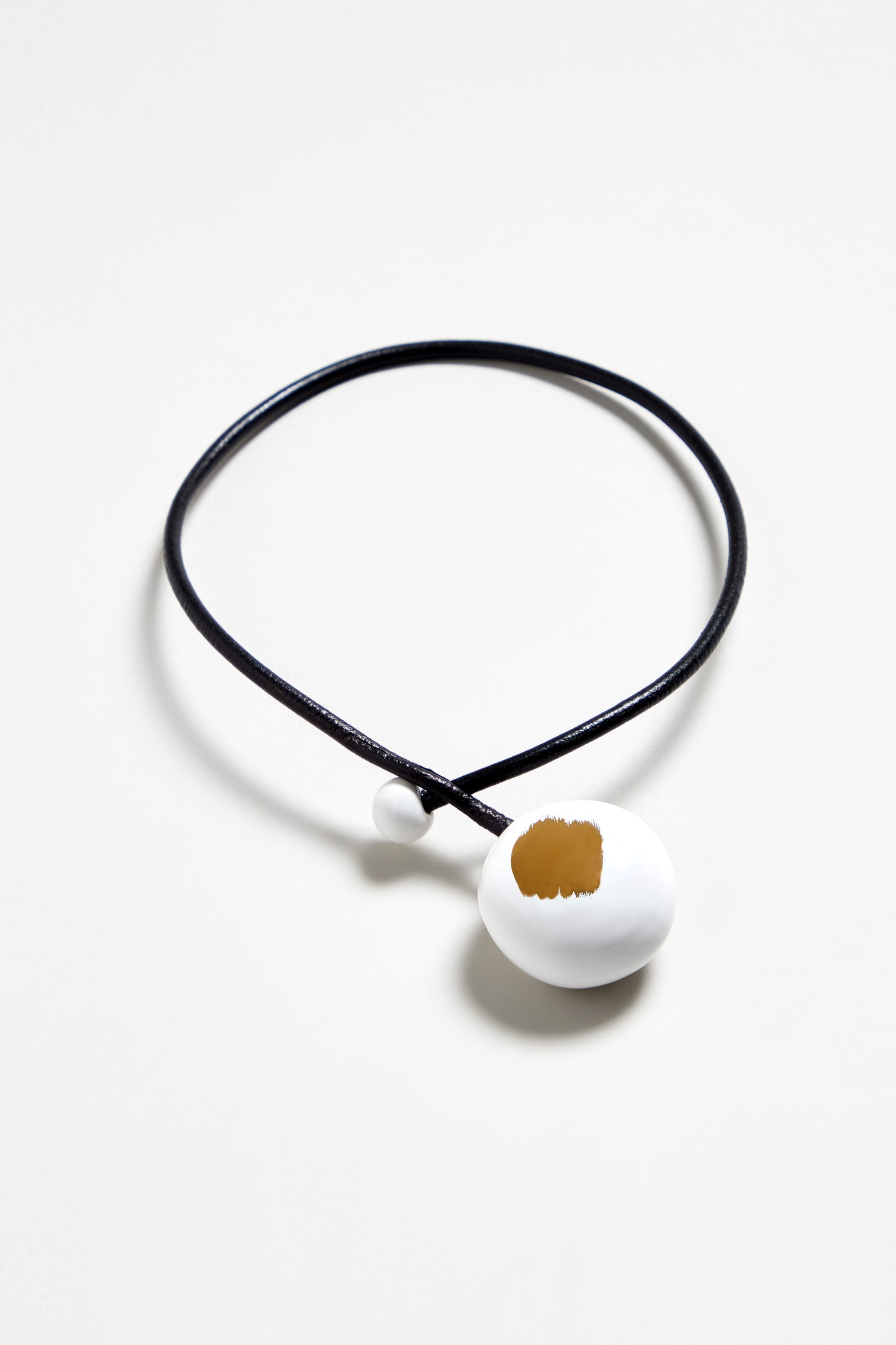 The Handmade Gold Brushstroke Ceramic Bead Necklace features a black cord and white round pendant, accented with gold brushstrokes. Expertly crafted as French handmade jewelry, it is displayed on a plain white background.