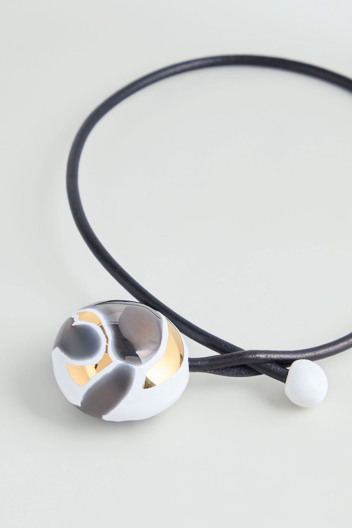 The Slate Handmade Ceramic Painted Bead Necklace features a handmade round pendant in white, gold, and gray with a black cord, inspired by French design. This artisan jewellery piece is displayed on a light background.