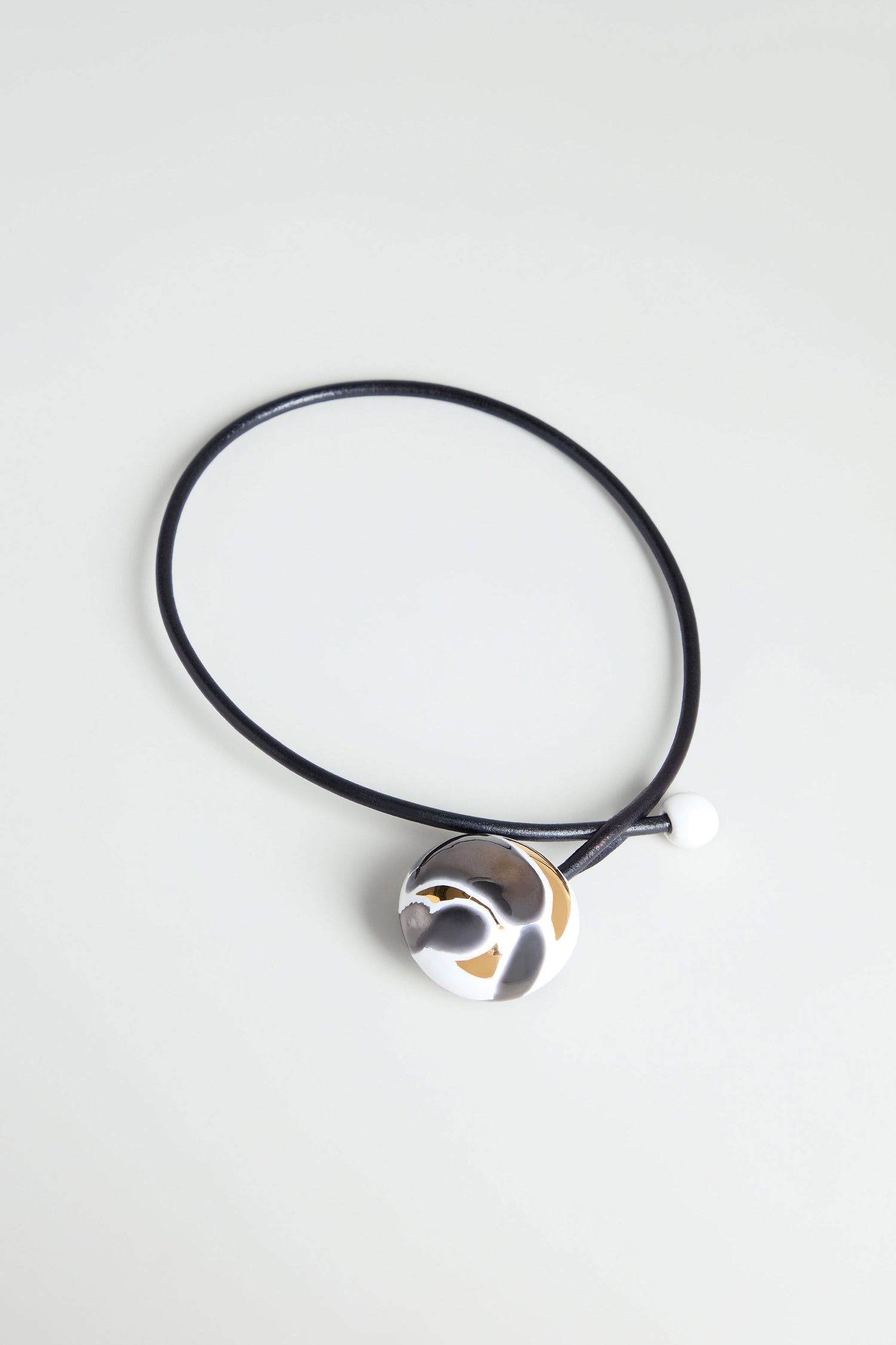 The Slate Handmade Ceramic Painted Bead Necklace features a black cord, metallic abstract pendant, and white ceramic bead—blending French-inspired elegance with artisan craftsmanship. Displayed on a light surface for a modern look.