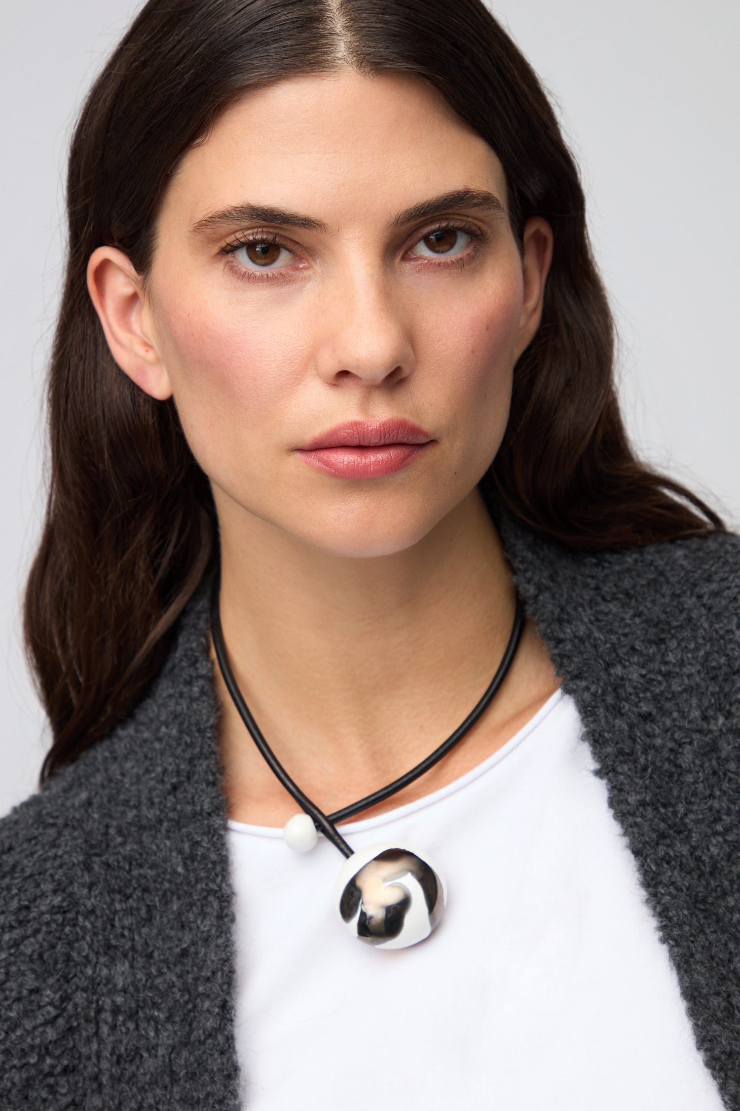 A woman with long dark hair wears a white top, gray textured cardigan, and the Slate Handmade Ceramic Painted Bead Necklace with a large round pendant—an elegant artisan piece—looking directly at the camera.