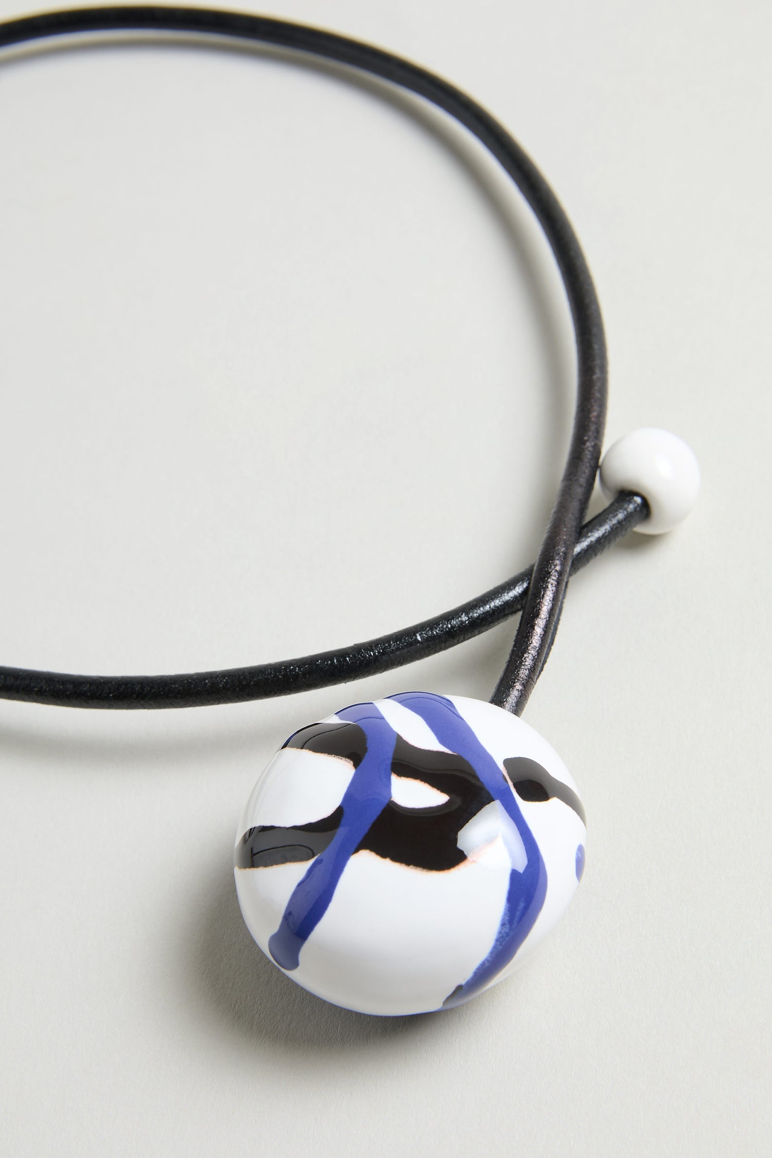 The Handmade Painted Ceramic Bead Necklace features a black leather cord, a round white pendant with blue and black abstract lines, and is accented by a small painted ceramic bead.