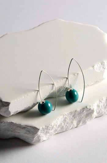 Handmade Ceramic Bead Earrings feature teal ceramic beads on silver wire with a sculptural twist, shown on stacked white stone slabs.