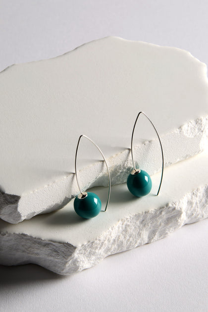 Handmade Ceramic Bead Earrings feature teal ceramic beads on silver wire with a sculptural twist, shown on stacked white stone slabs.