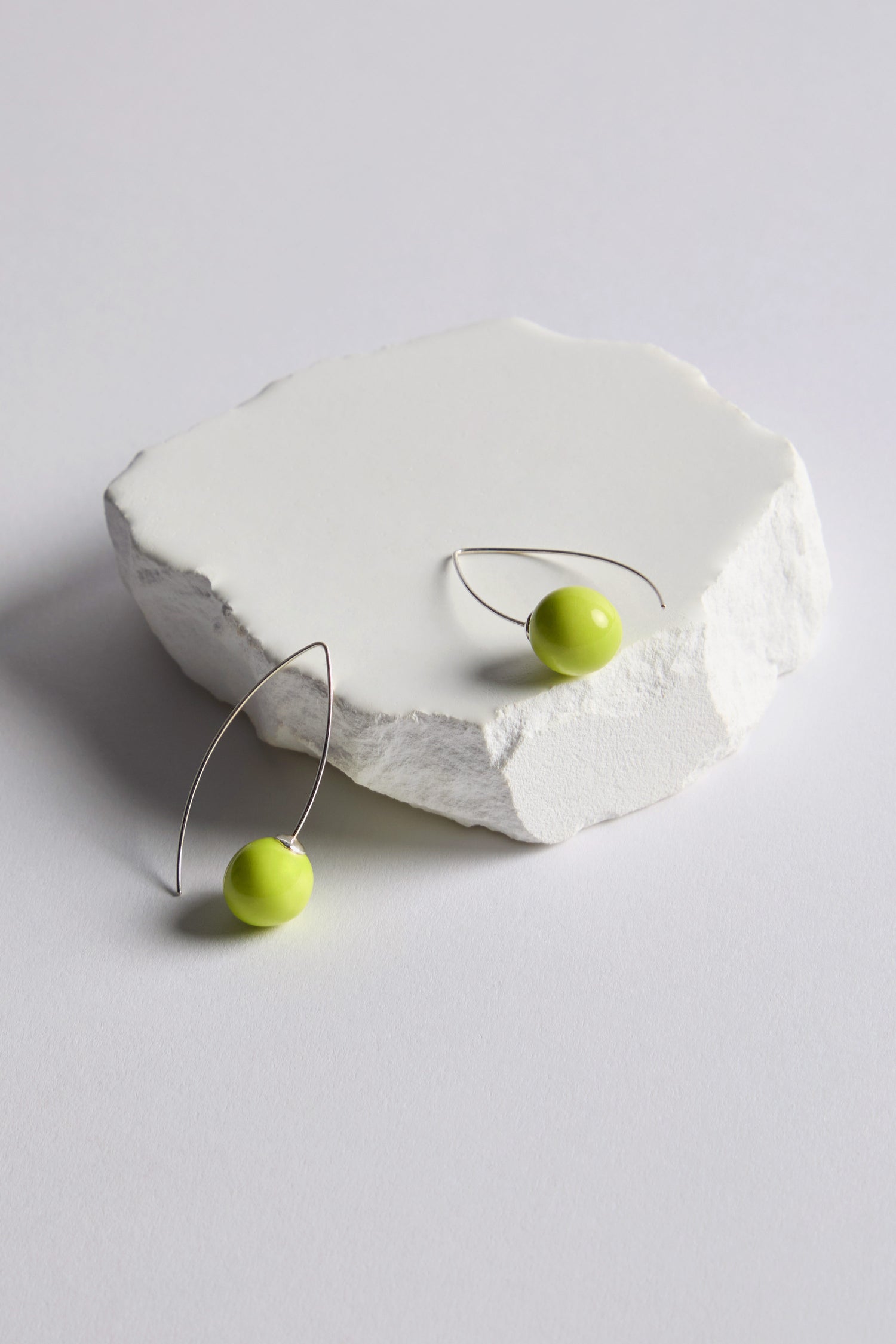 Handmade Ceramic Bead Earrings featuring two silver wire earrings with round, bright yellow-green ceramic beads and an organic finish are displayed on a white textured stone surface against a plain background.