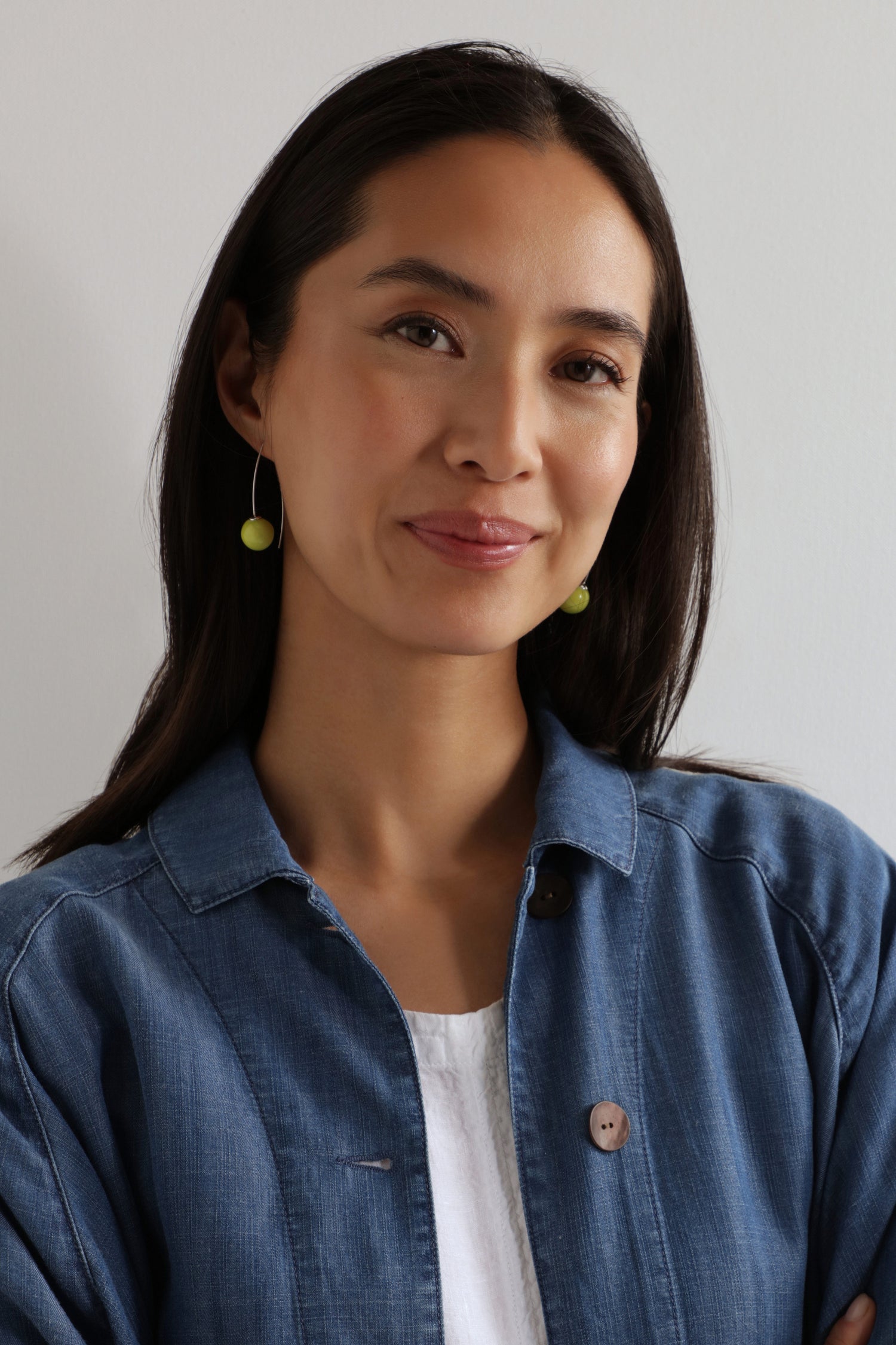 A woman with long dark hair wears a blue button-up shirt over a white top and the Handmade Ceramic Bead Earrings, standing against a plain light-colored wall.