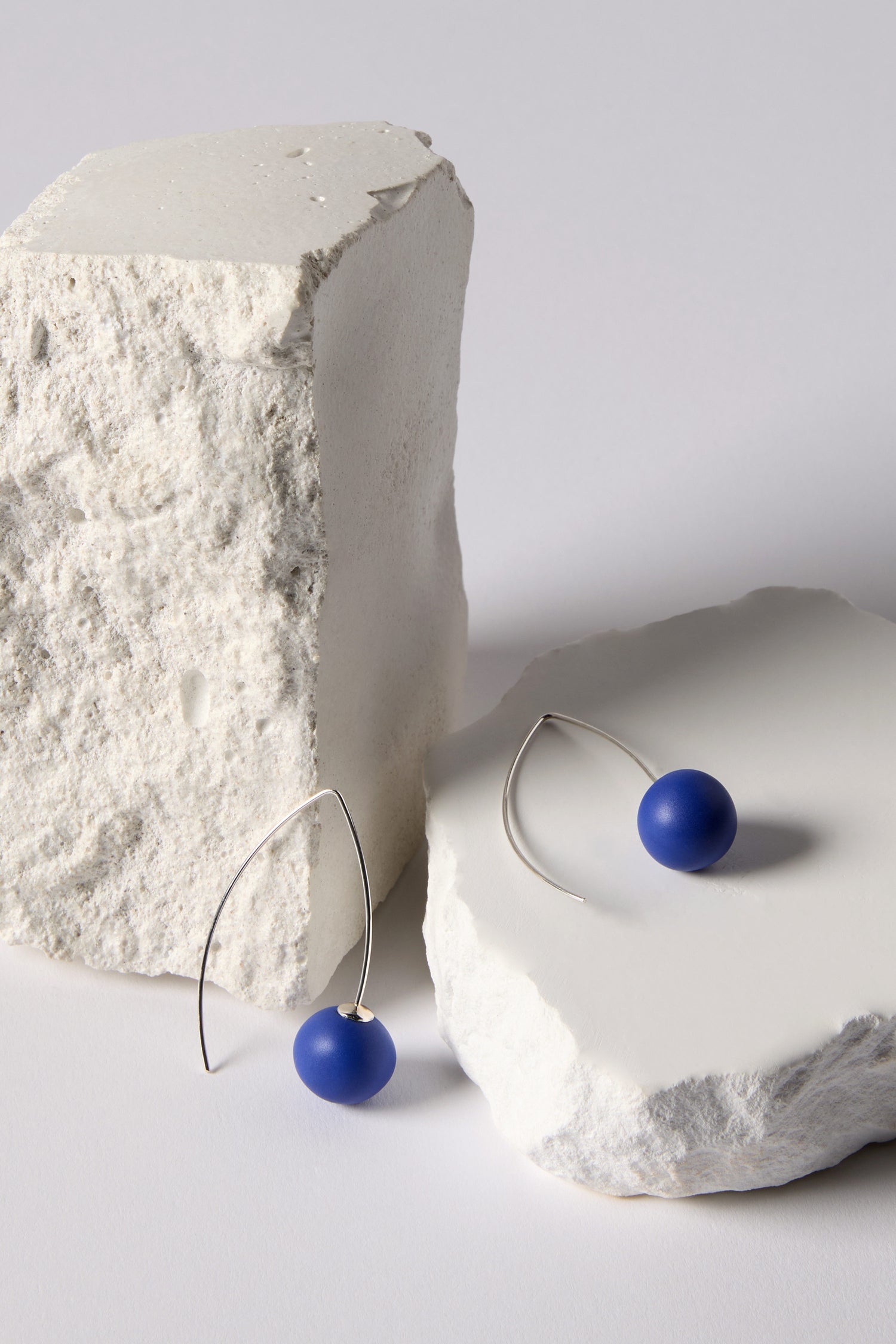 Handmade Ceramic Bead Earrings with blue spheres and an organic finish, shown on two rough white stone blocks.