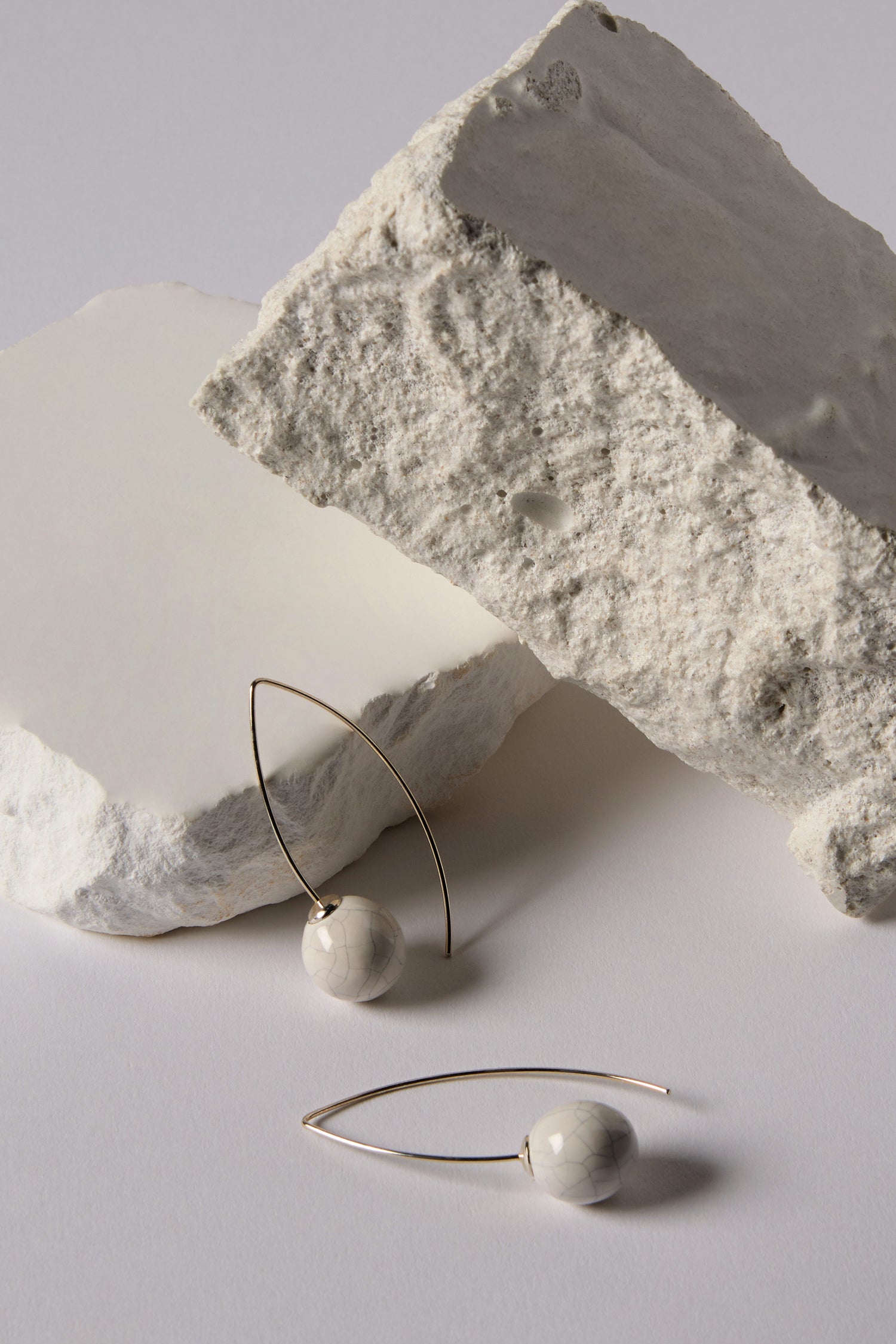 Handmade Ceramic Bead Earrings with white marbled spheres and thin metal hoops are showcased on a white surface next to rough white stone blocks, emphasizing their organic finish.