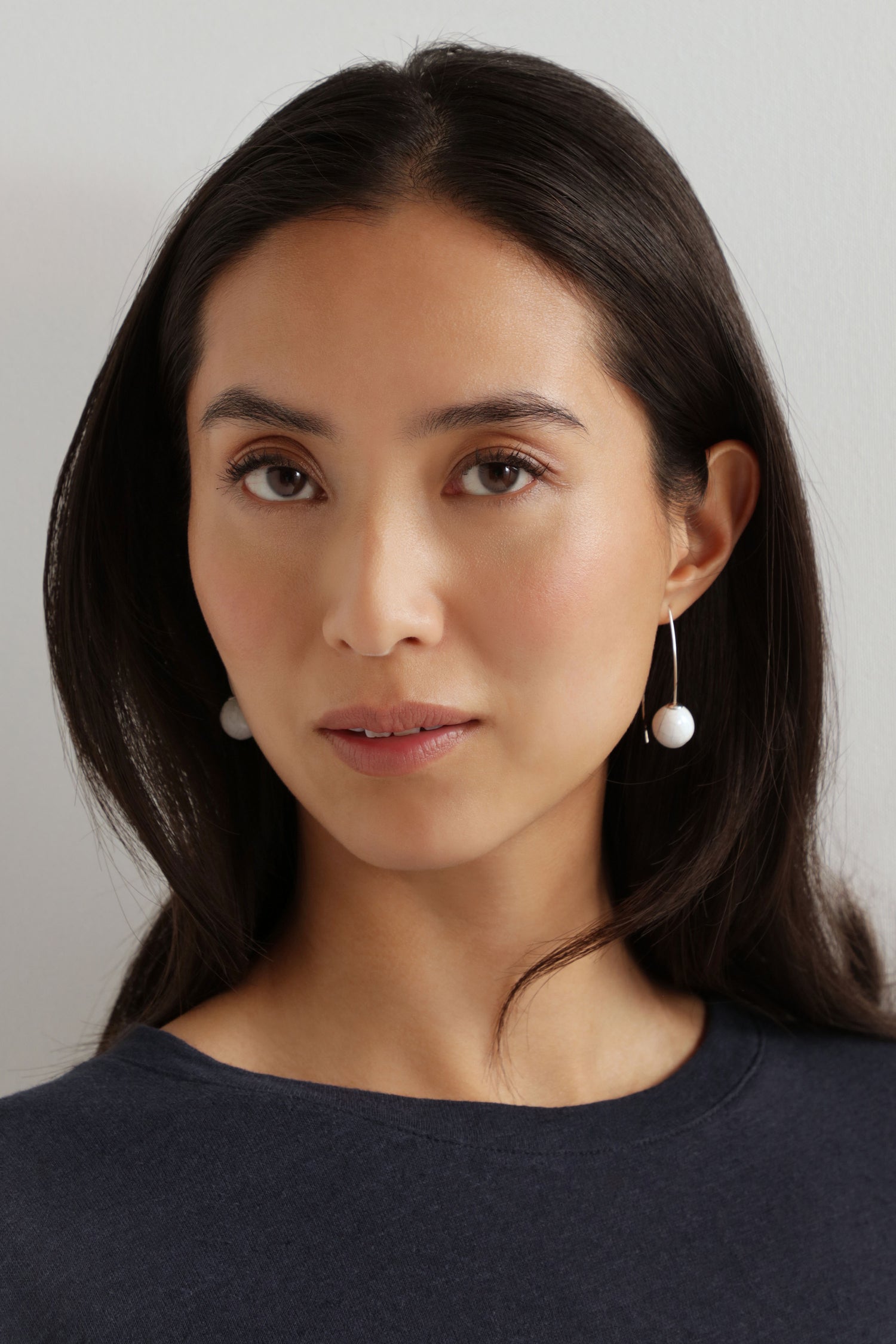 A woman with long dark hair wears a navy top and Handmade Ceramic Bead Earrings with an organic finish, gazing at the camera against a plain light background.