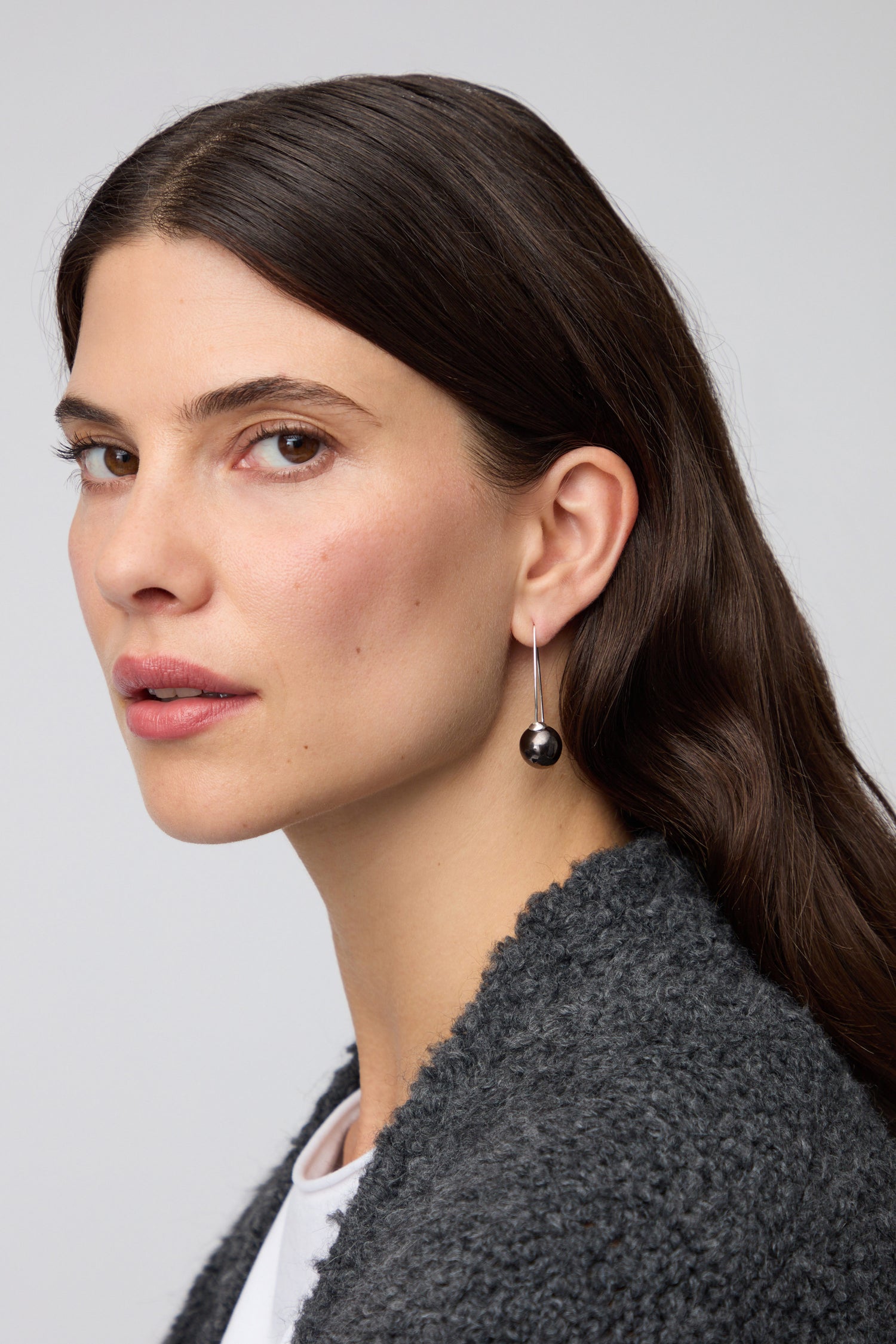 A woman with long brown hair wears a dark textured jacket and a single Handmade Ceramic Graphic Metal Earring as she looks toward the camera against a plain background.
