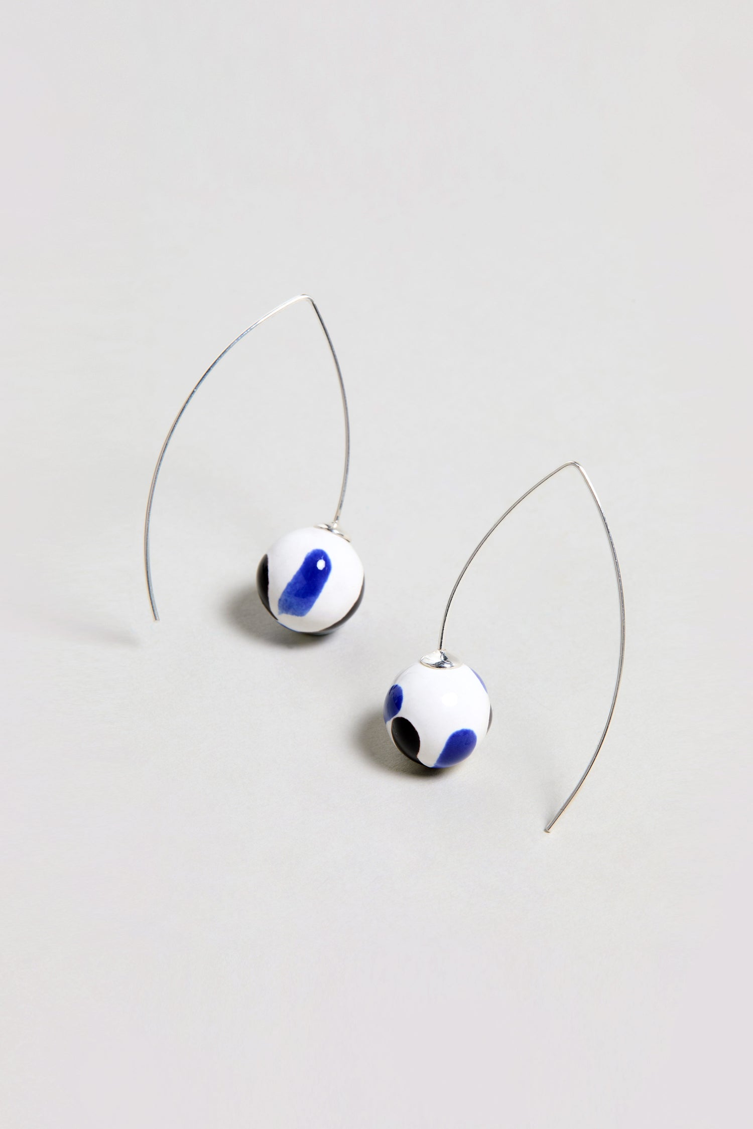 Handmade Painted Ceramic Bead Earrings feature white ceramic spheres with blue and black abstract spots, each elegantly suspended from a curved silver wire.