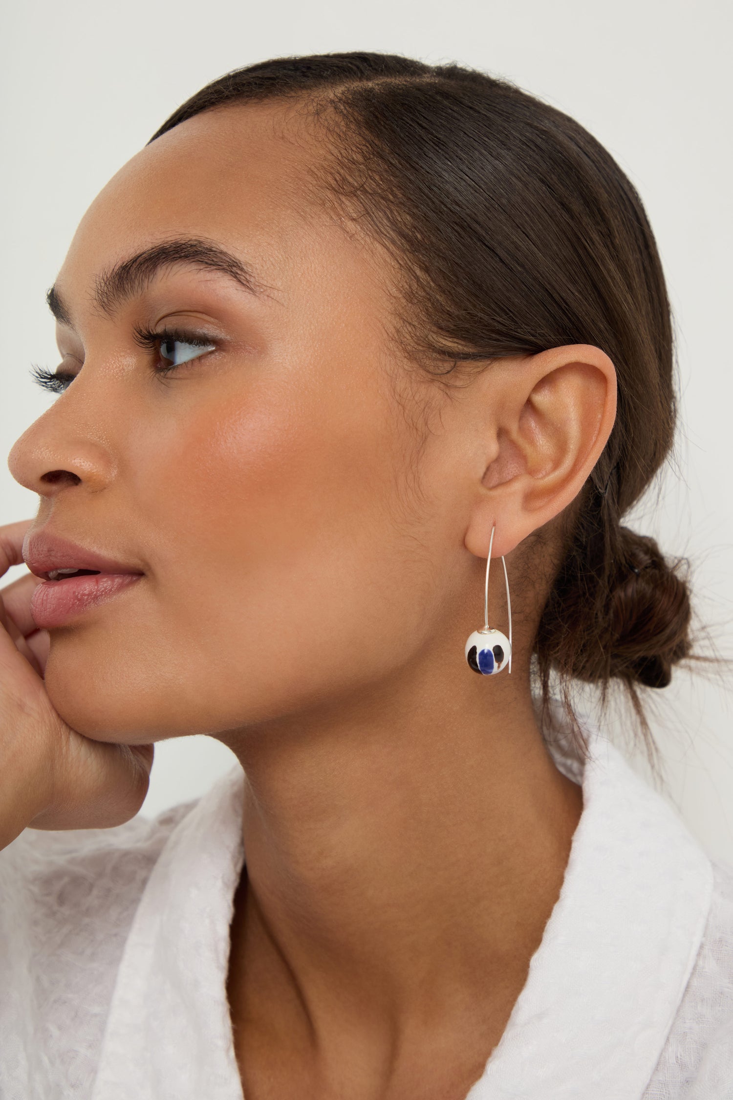 Woman with smooth skin and tied-back hair wears Handmade Painted Ceramic Bead Earrings—silver hooks with blue and white ceramic beads—paired with a white textured top, resting her chin on her hand and facing left.