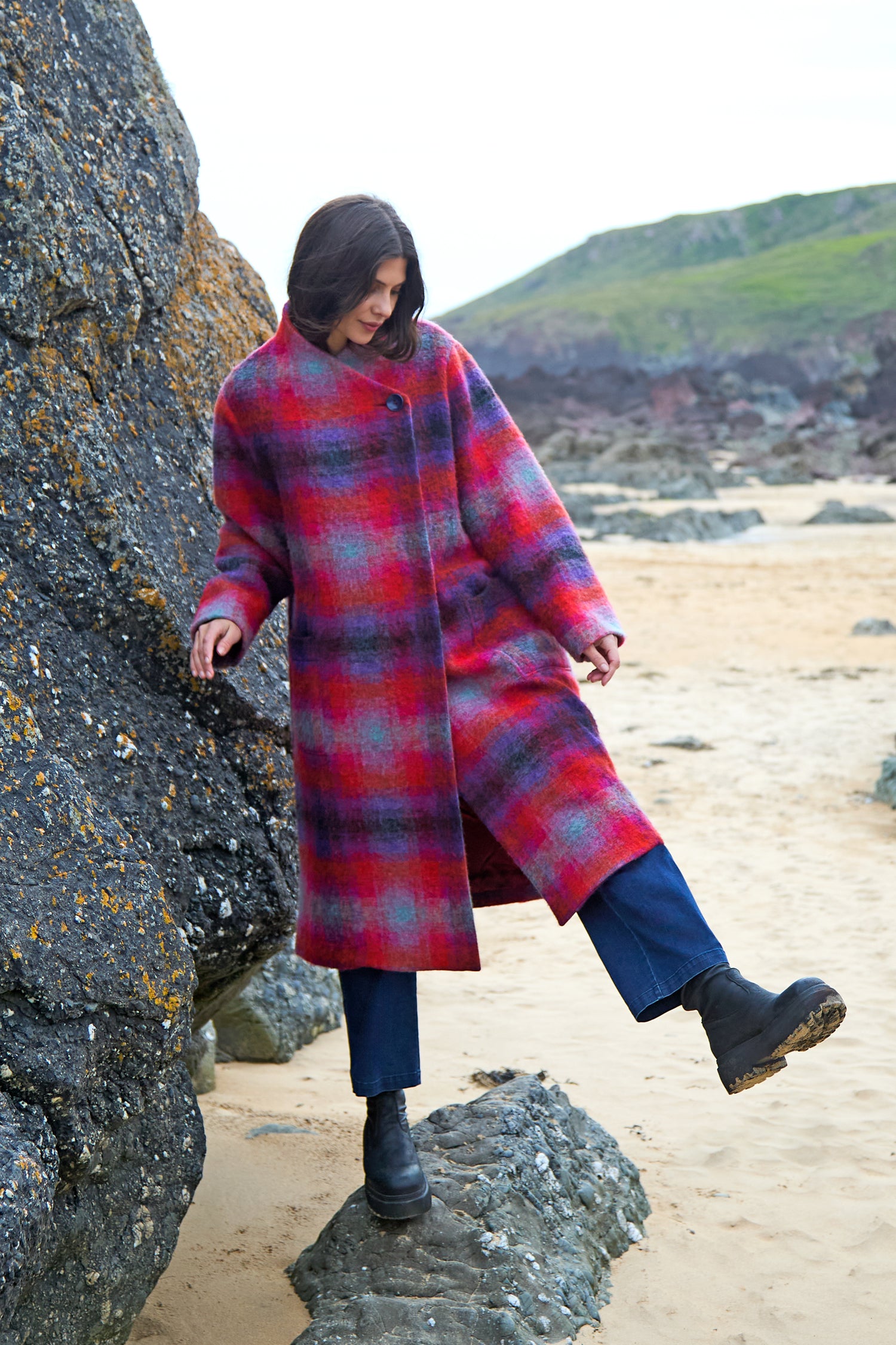 Wearing the Bright Brushed Check Wool Coat and black boots, a person stands on rocks at a sandy beach with grassy hills in the background.