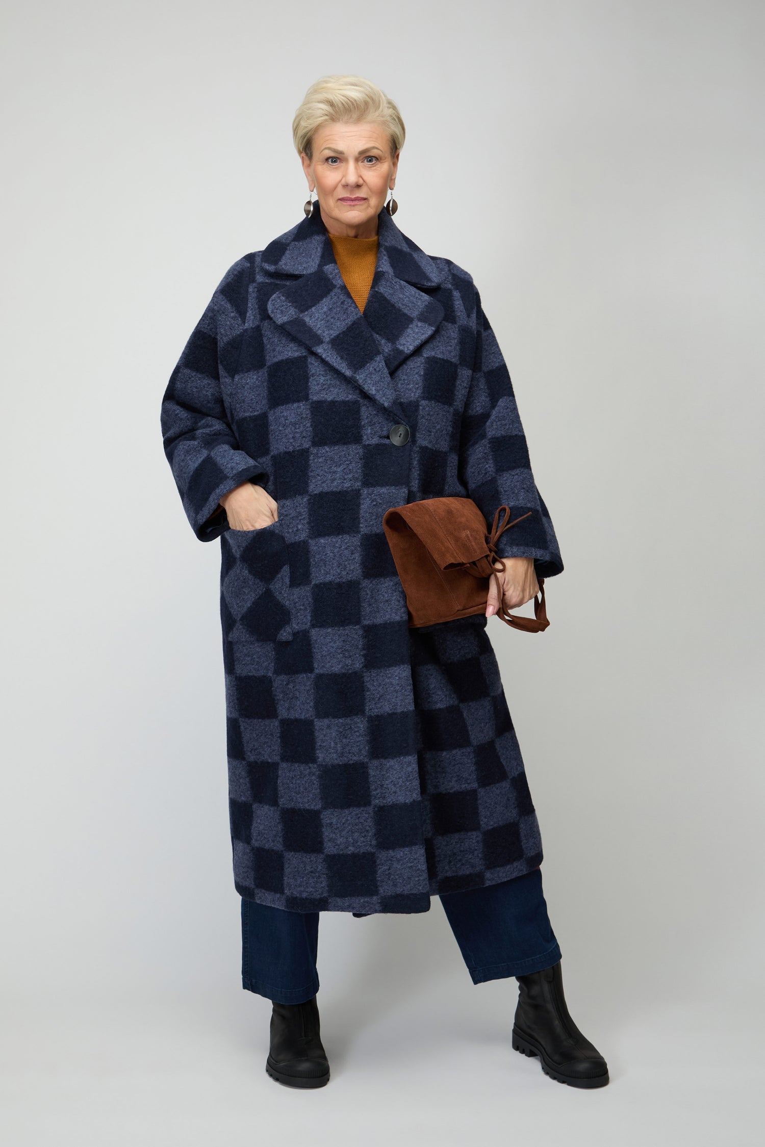 An older woman wears the Checkerboard Wool Coat in blue, paired with blue pants and black boots, holding a brown bag against a plain light background.