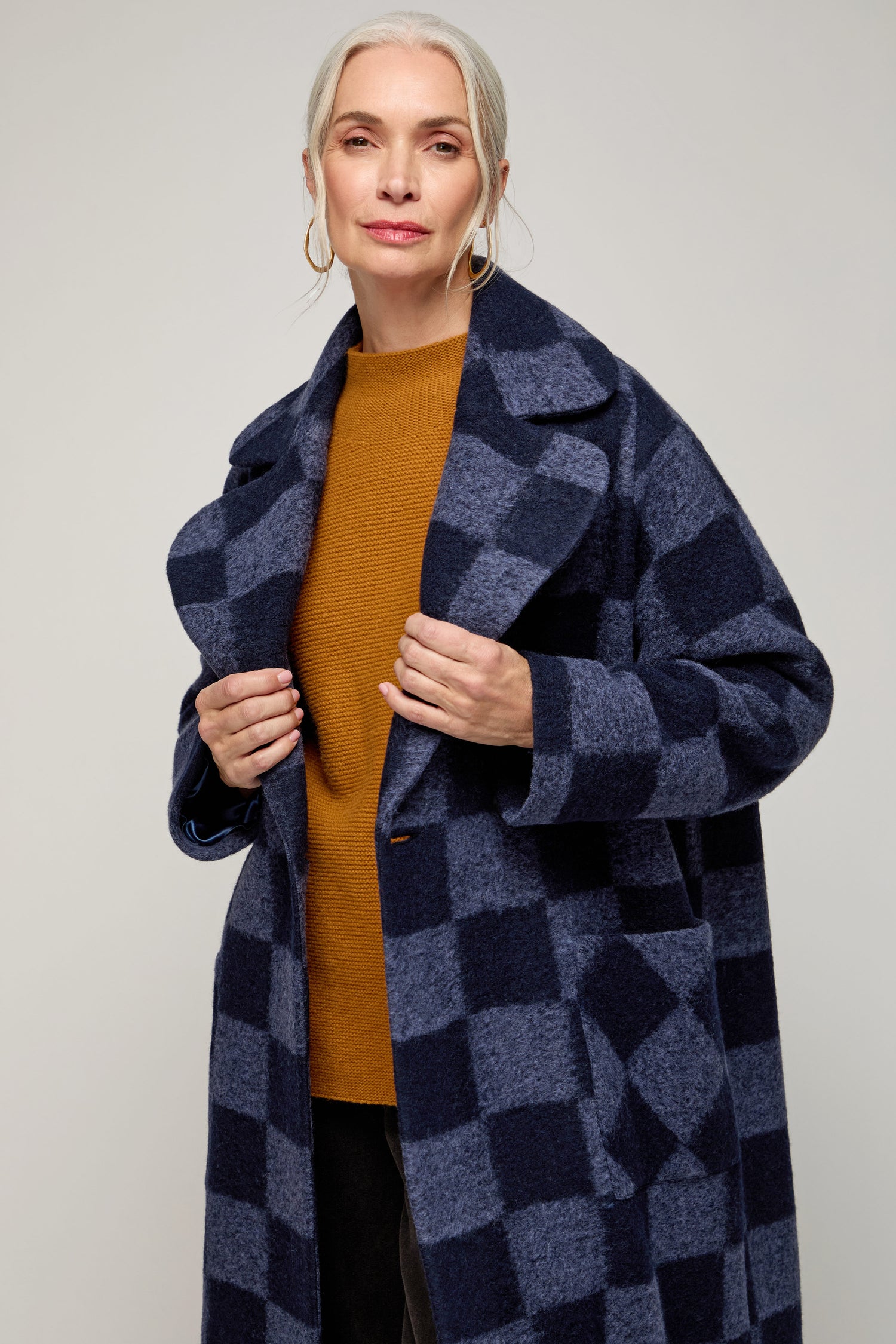 A woman with gray hair wears the Checkerboard Wool Coat over a mustard sweater, standing against a plain background.
