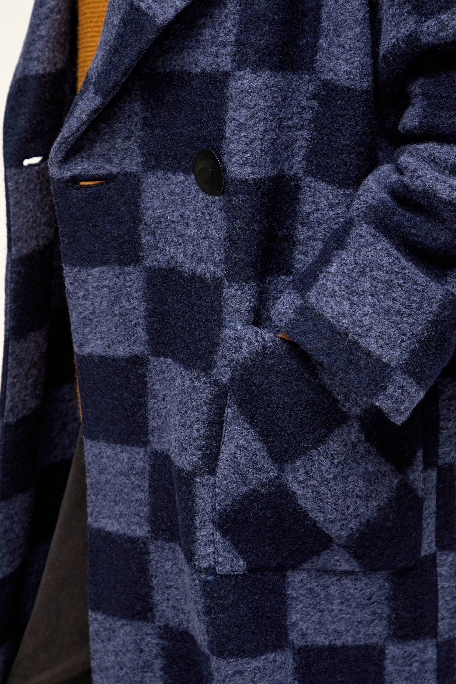 Close-up of a person wearing the Checkerboard Wool Coat, a British-designed coat in navy and blue with a large black button, shown with their hand in the side pocket.