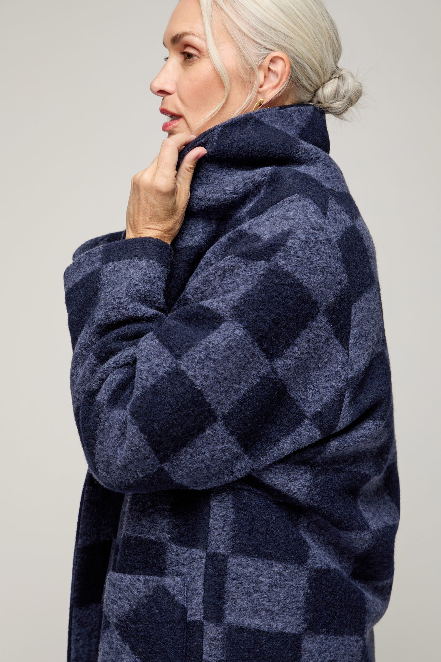An older woman with light hair in a low bun wears the Checkerboard Wool Coat, holding its collar close to her face. The coat's British design stands out in profile against a plain background.