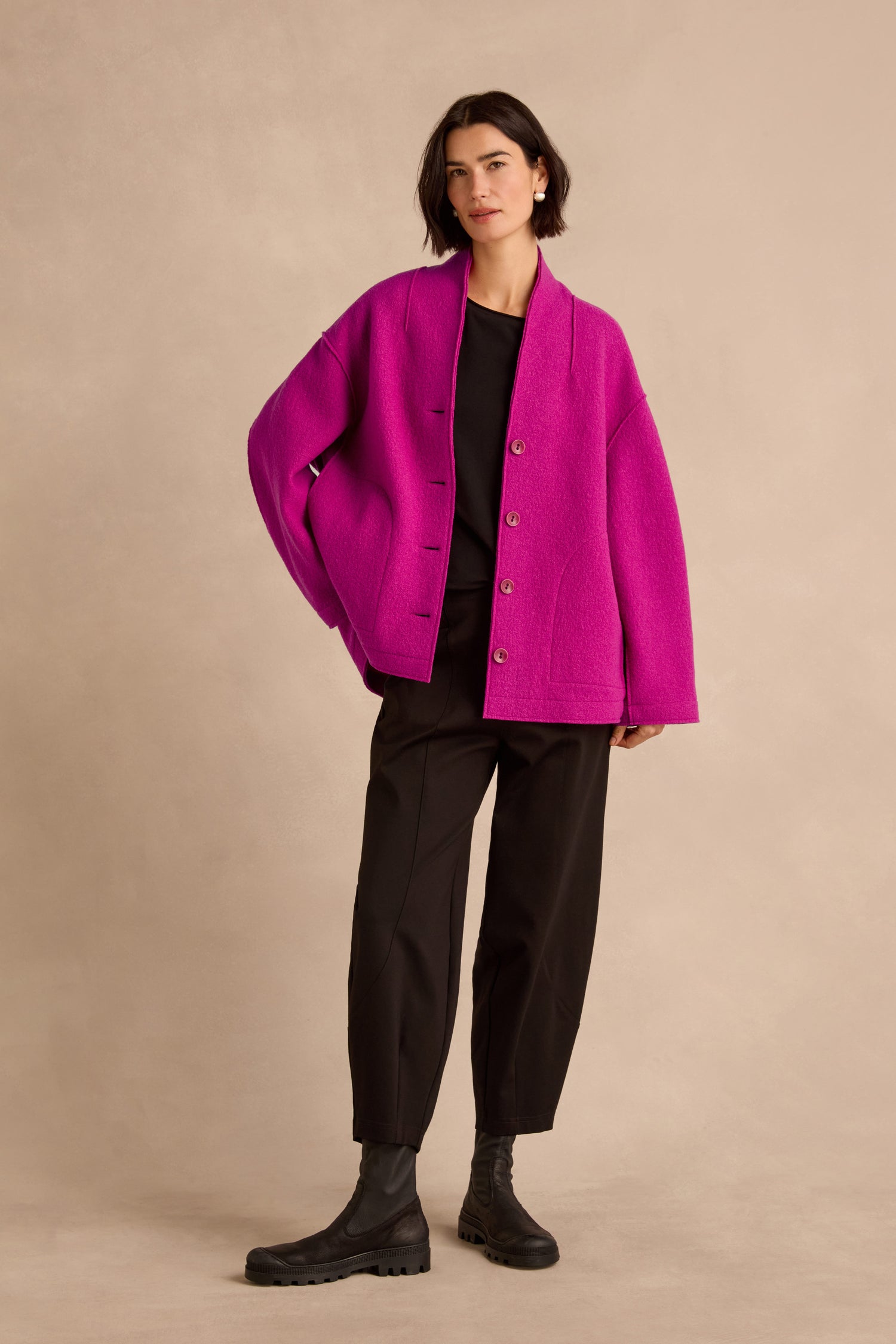 Person standing against a beige background wearing the Magenta Boiled Wool Cocoon Jacket, styled with a black shirt, black pants, and black boots.
