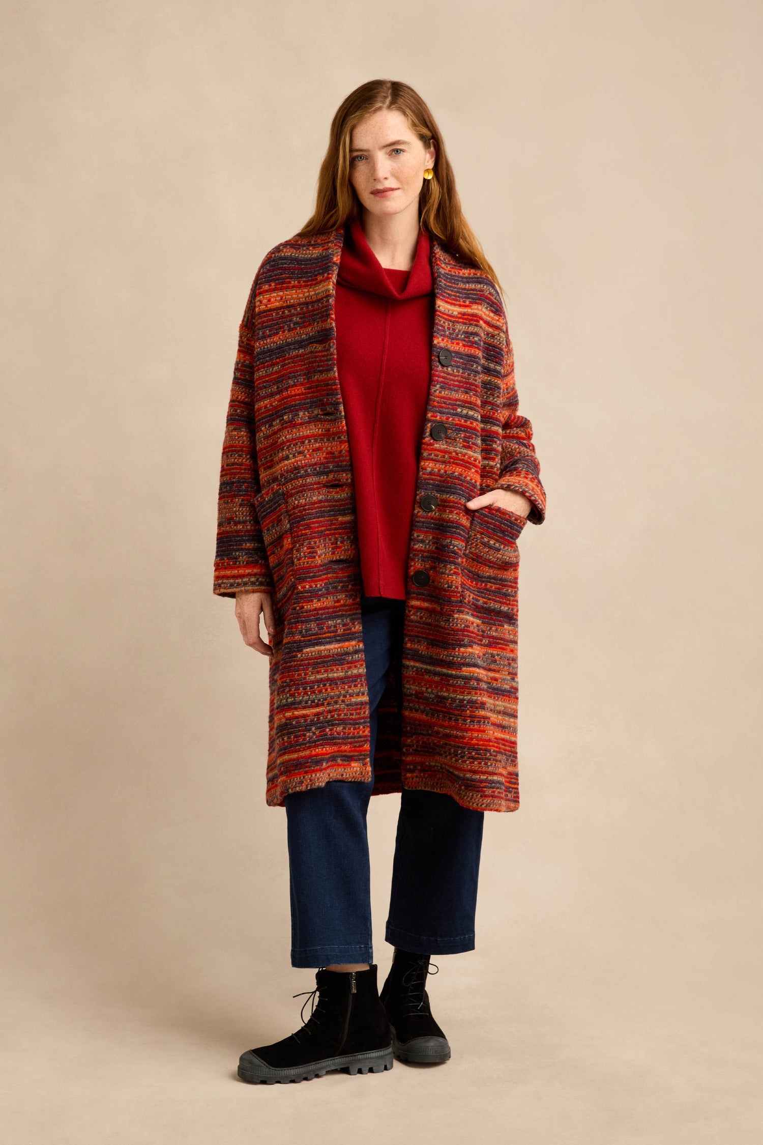 A person stands against a neutral background wearing the Artisan Wool Blend Stripe Coat with a red patterned design, layered over a red top, paired with dark blue pants and black boots.