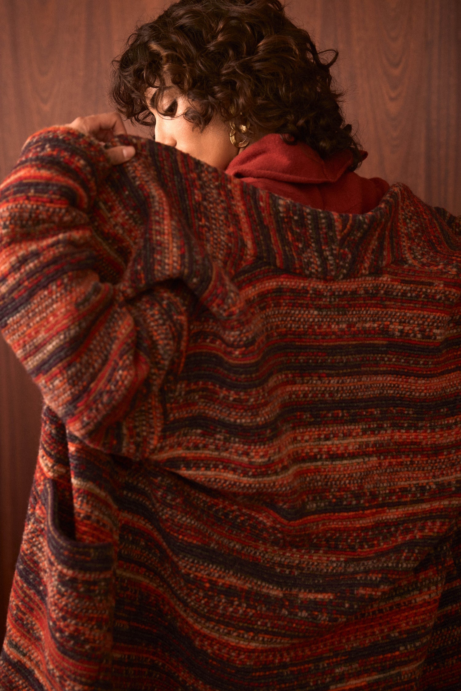 A person with curly hair is turned away from the camera, partially covering their face, as they stand in front of a wooden background wearing the oversized Artisan Wool Blend Stripe Coat featuring bold red and brown patterns.