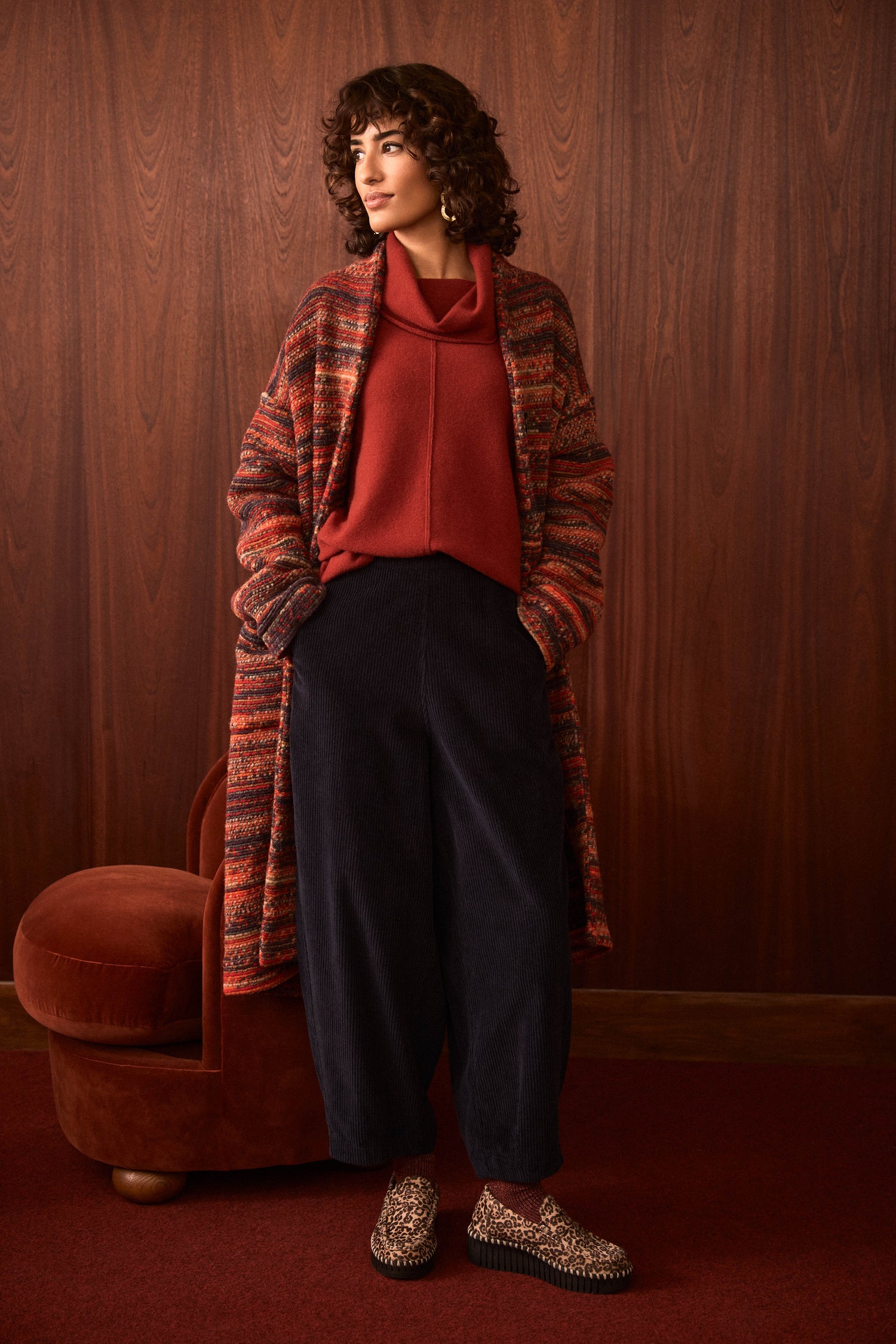 A woman stands indoors against a wood-paneled wall, wearing a red sweater, dark pants, leopard-print shoes, and the Artisan Wool Blend Stripe Coat layered on top; her hands are in her pockets as she looks to the side.