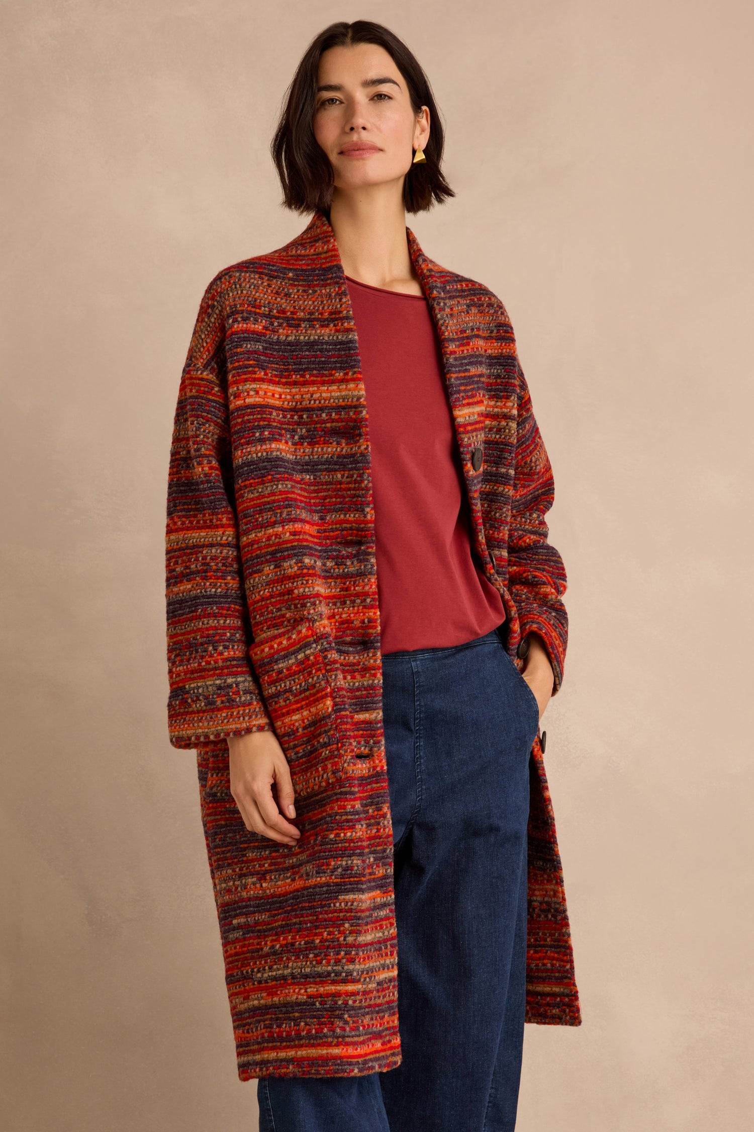 A woman models the Artisan Wool Blend Stripe Coat—a long, open-front coat featuring red, orange, and gray stripes—layered over a red shirt and blue pants against a beige background.