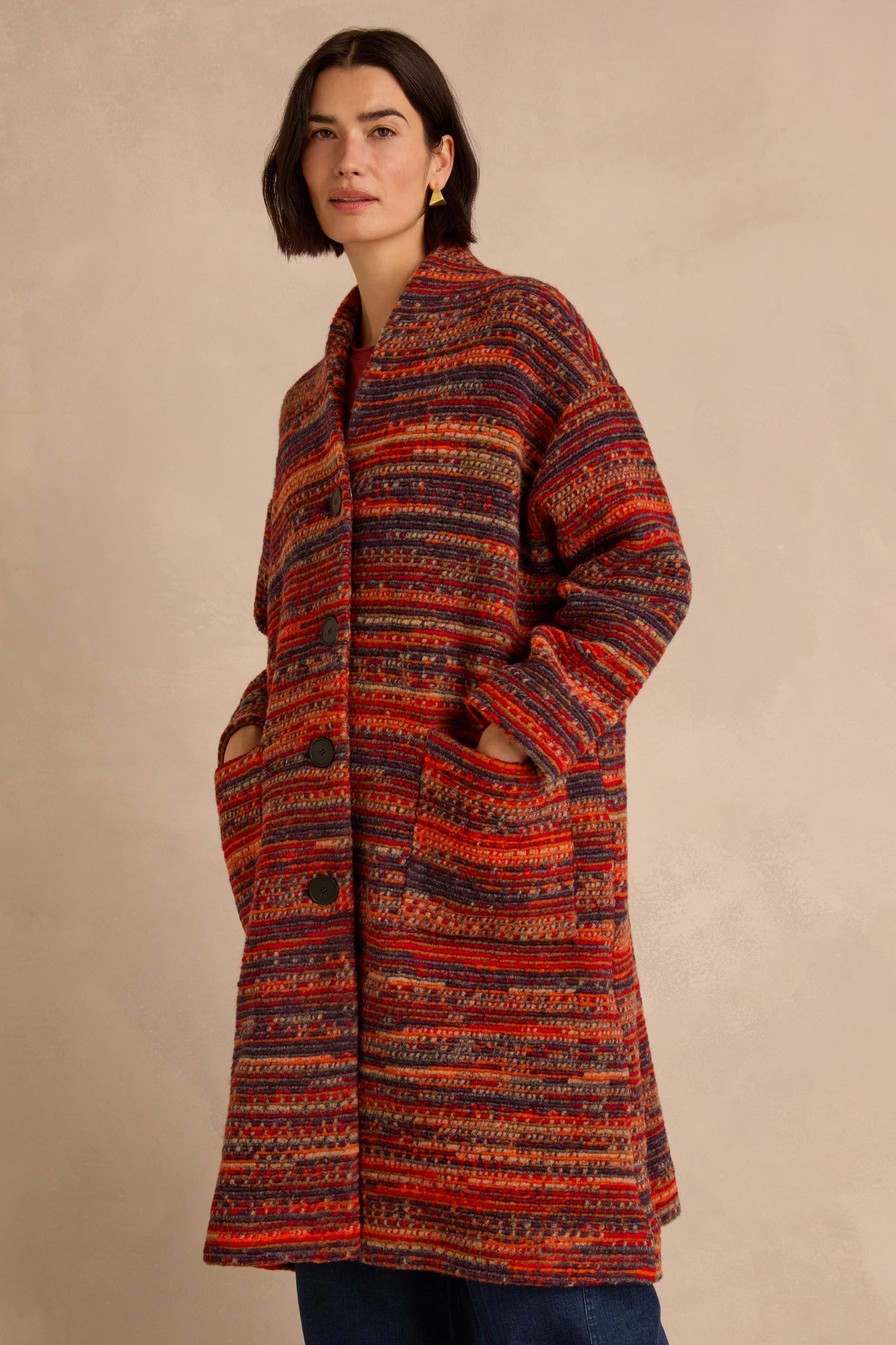 A woman stands against a neutral background wearing the Artisan Wool Blend Stripe Coat, a loose, knee-length piece featuring vibrant red and orange stripes and large pockets.