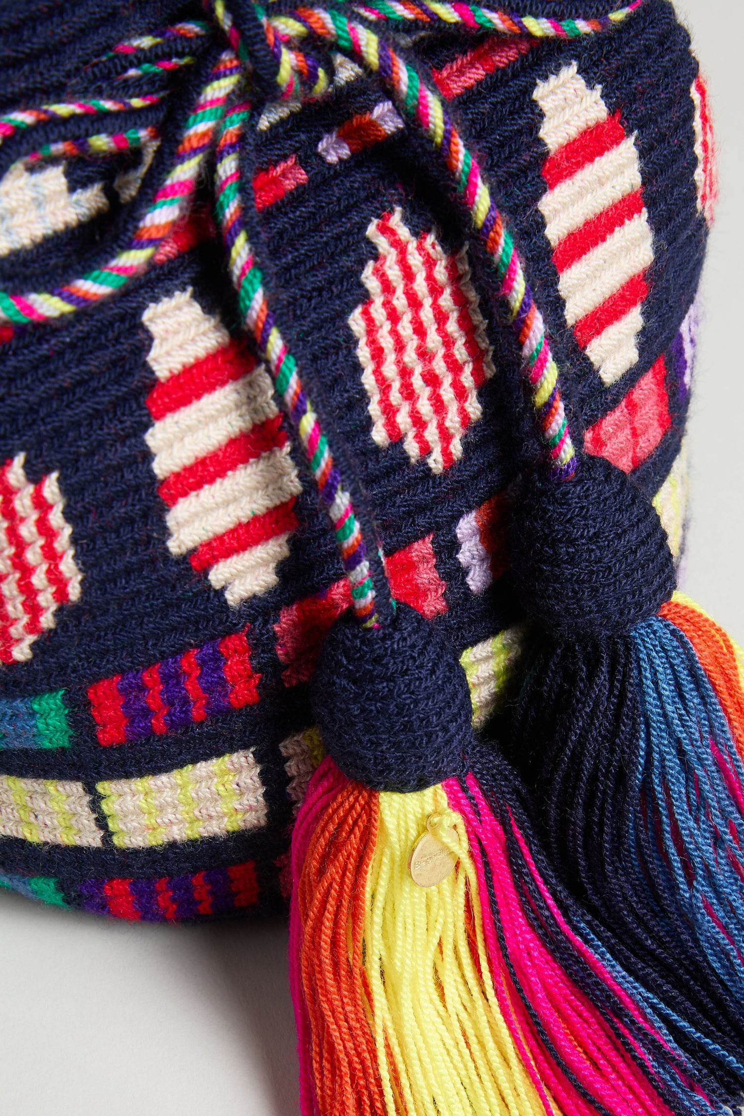 A close-up of the Hand Crocheted Wayuu Geometric Crossbody Bag in navy blue with red, white, and multicolored geometric patterns, accented by twisted drawstrings and large colorful tassels.