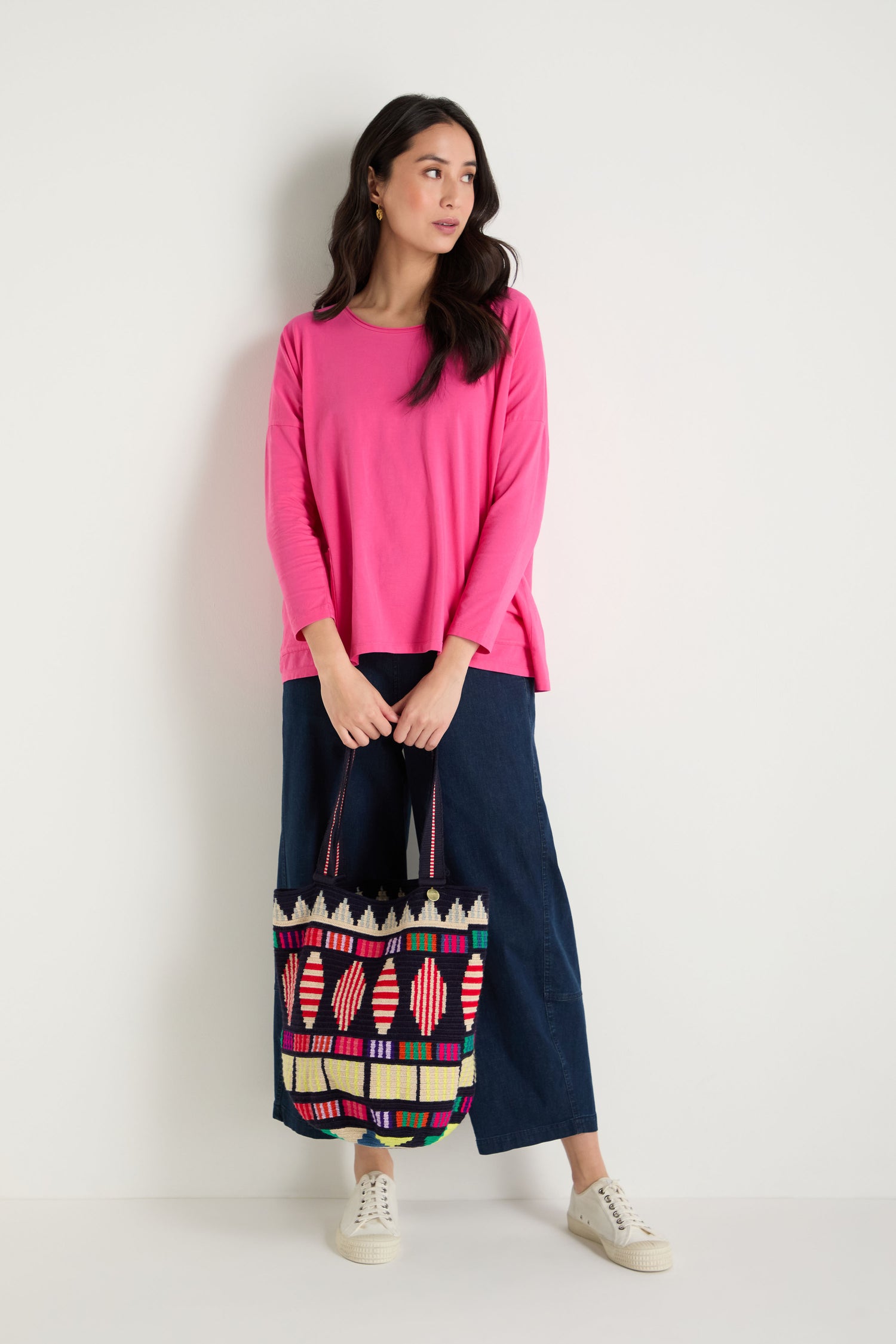 Woman stands against a plain wall wearing a bright pink long-sleeve top, wide-leg blue pants, white sneakers, and holding the Hand Crocheted Cala Geometric Tote Bag.