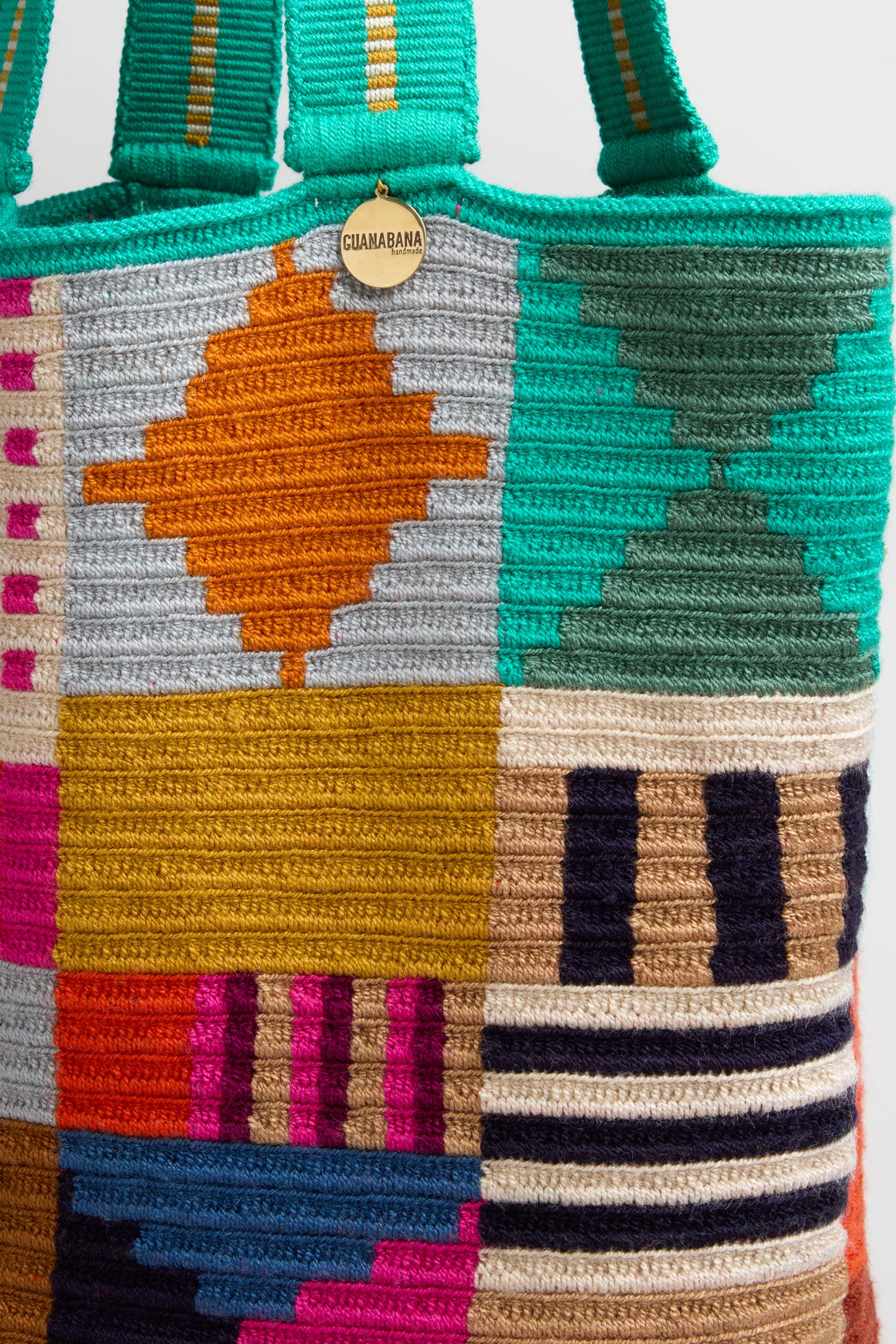 The Hand Crocheted Cala Tote Bag features a colorful geometric design in orange, teal, navy, pink, and beige, and includes a small round "GUANABANA" tag near the top.